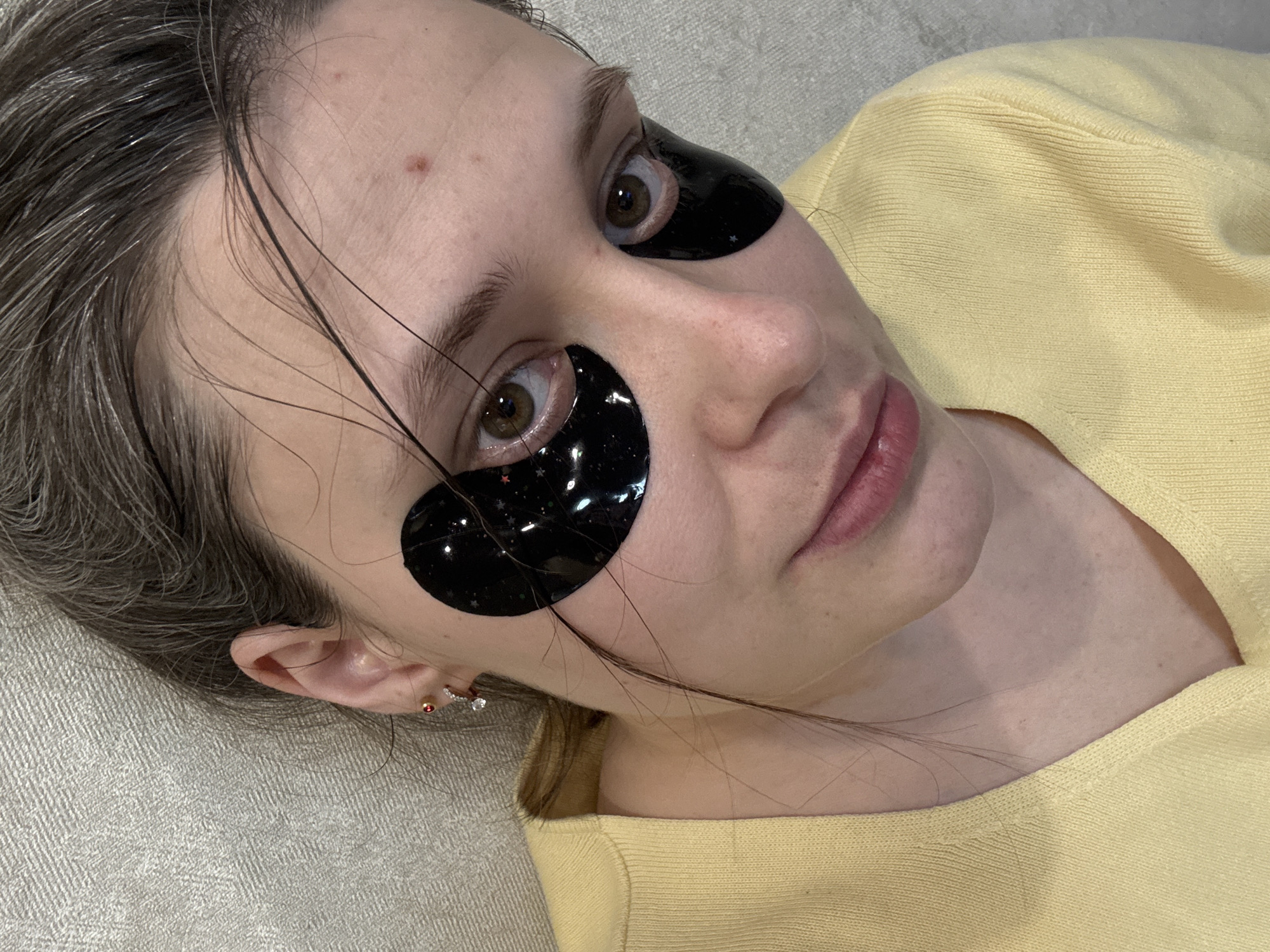 A woman lies on a light grey textured surface, possibly fabric or upholstery, wearing a yellow top. She has black under-eye patches with small star patterns on them. Her hair is greyish-brown and slightly wet, with some strands falling across her face. She has fair skin and light brown eyes. The woman is wearing multiple earrings in her left ear. The location is not specified or discernible from the image; the description requested includes "Chaikovsky, Russia" which is not visibly connected to the content.