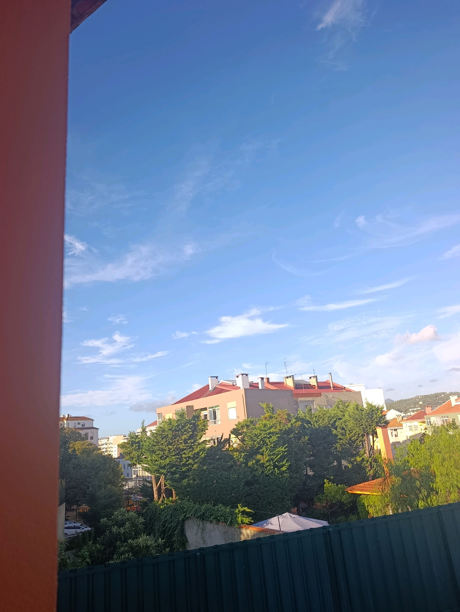 The image depicts a view of rooftops and trees against a bright blue sky with scattered cirrus clouds. The foreground is dominated by a reddish-orange structure on the left, partially obscuring the scene. In the middle ground, several multi-story buildings with reddish-tiled roofs are visible, interspersed with lush green trees. Some buildings have visible chimneys and antennas. A dark green fence runs along the bottom of the frame, obscuring the ground level. A white umbrella structure is visible peeking out from behind some trees. The overall scene suggests an urban environment in a residential area. Based on the architectural style of the buildings with their red-tiled roofs and the presence of Mediterranean vegetation, this scene is likely located in Setúbal, Portugal. There are no people or active events depicted in the image.