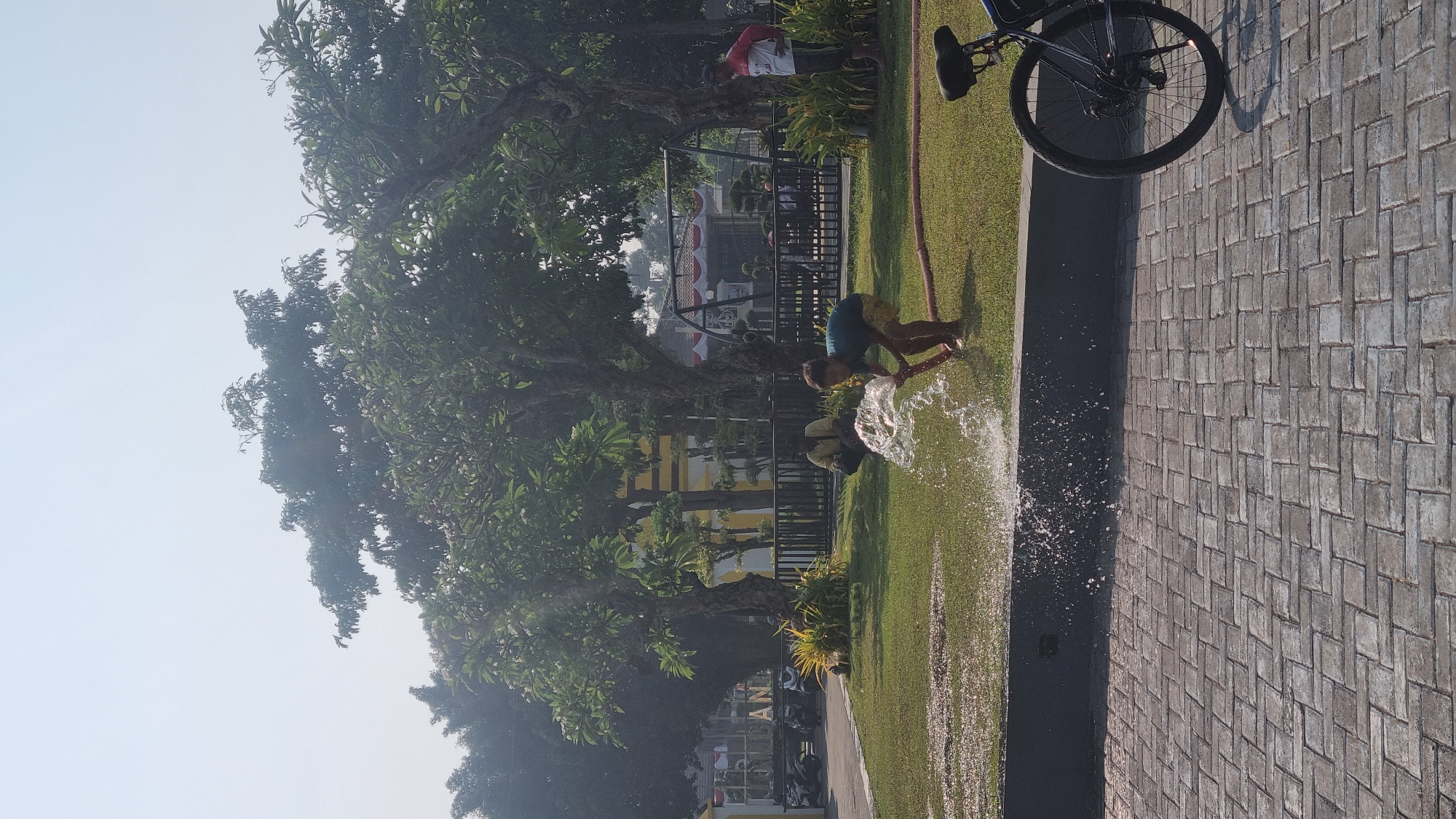 In Jombang, Indonesia, a young person waters the grass with a hose. The scene takes place outdoors in a park-like area with large trees and manicured lawns. A paved walkway made of grey interlocking bricks is in the foreground. To the right, a black bicycle is parked. In the background, a fence encloses a playground or recreational area with a swing set visible. Several people are present, including one person in a red and white shirt standing, another person in a yellow shirt crouching, and two other individuals partially obscured behind the fence. The sky is bright and hazy, suggesting a sunny day.