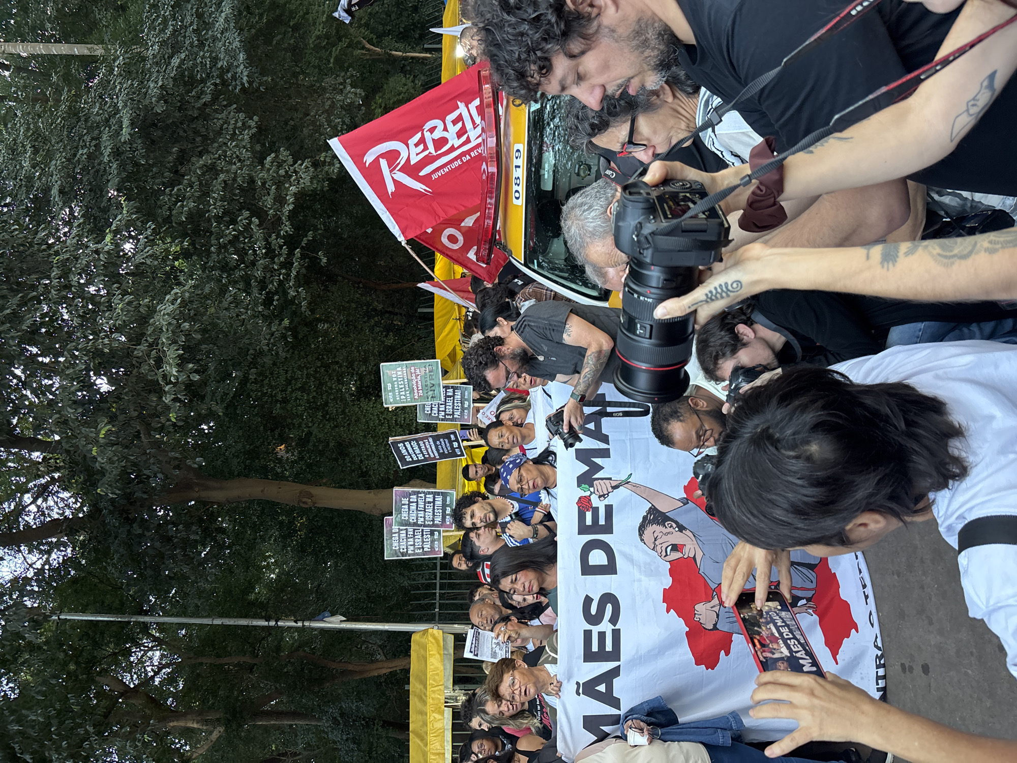 The image shows a rally in the city of São Paulo, Brazil. A group of people, some holding signs, are displaying a banner that reads "Mães de Maio" (Mothers of May). Several photographers are documenting the event. One person is taking a photo of the banner with their phone. There is a red flag with the word "Rebelde" (Rebel) on it and a yellow bus behind the crowd. The environment includes trees and an urban setting. The signs held by the protestors have phrases such as "Chega de Chacina PM na Favela" (Stop the Slaughter PM in the Favela) and references to Palestine and Israel.