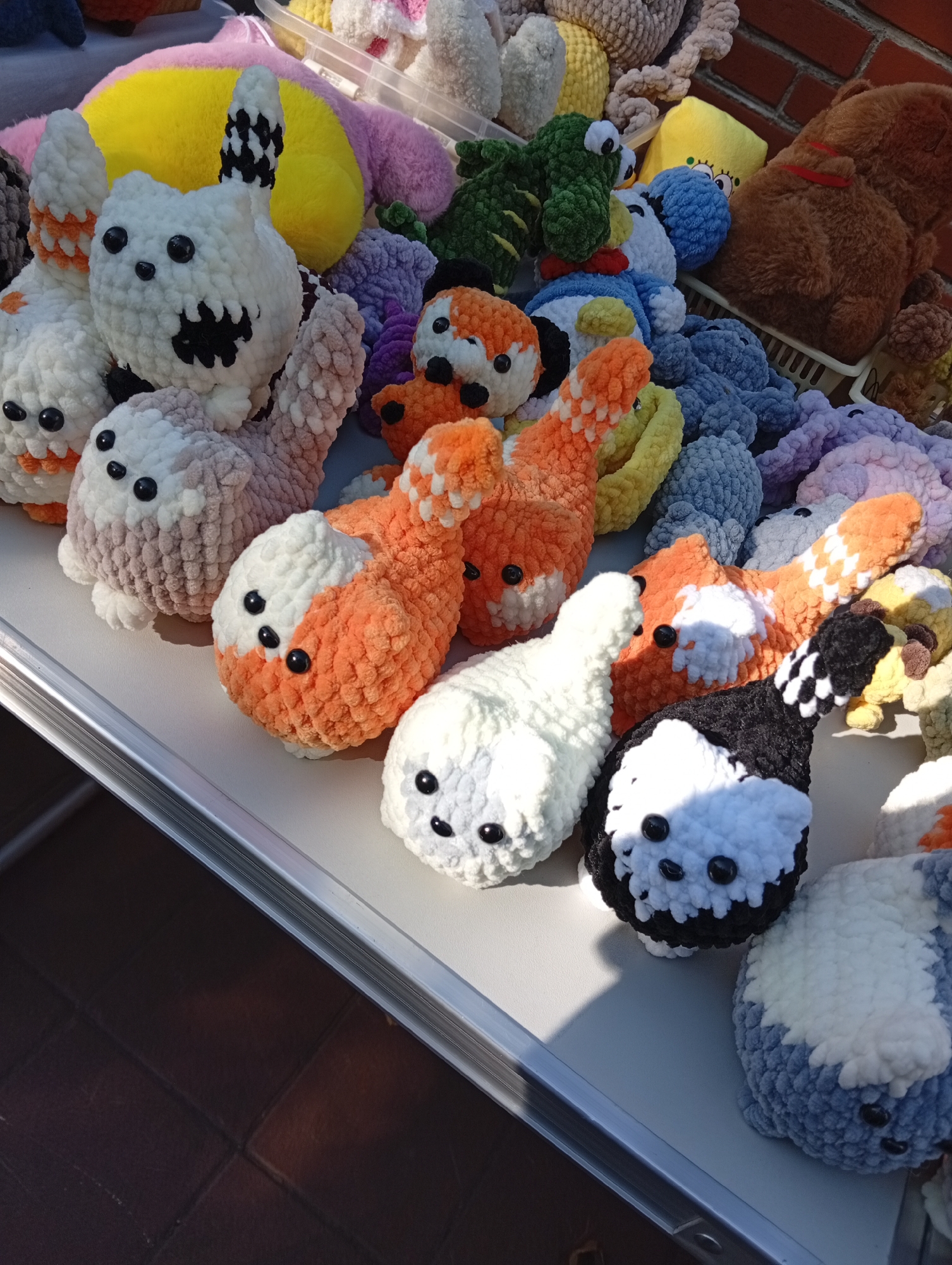 A close-up, eye-level, outdoor shot shows a collection of crocheted stuffed animals displayed on a white table. The stuffed animals are various colors and designs, including foxes, cats, and other creatures. Some are white with orange markings, others are white with black markings, and some are solid colors like orange, pink, purple, and blue. The animals have black button eyes and crocheted mouths. In the background, a brick wall is visible, suggesting an outdoor market or vendor stall. The lighting is natural and appears to be from sunlight, casting shadows on the table. The image is focused on the stuffed animals, with the background slightly blurred. The location is identified as a street market in Kostanay, Kazakhstan.