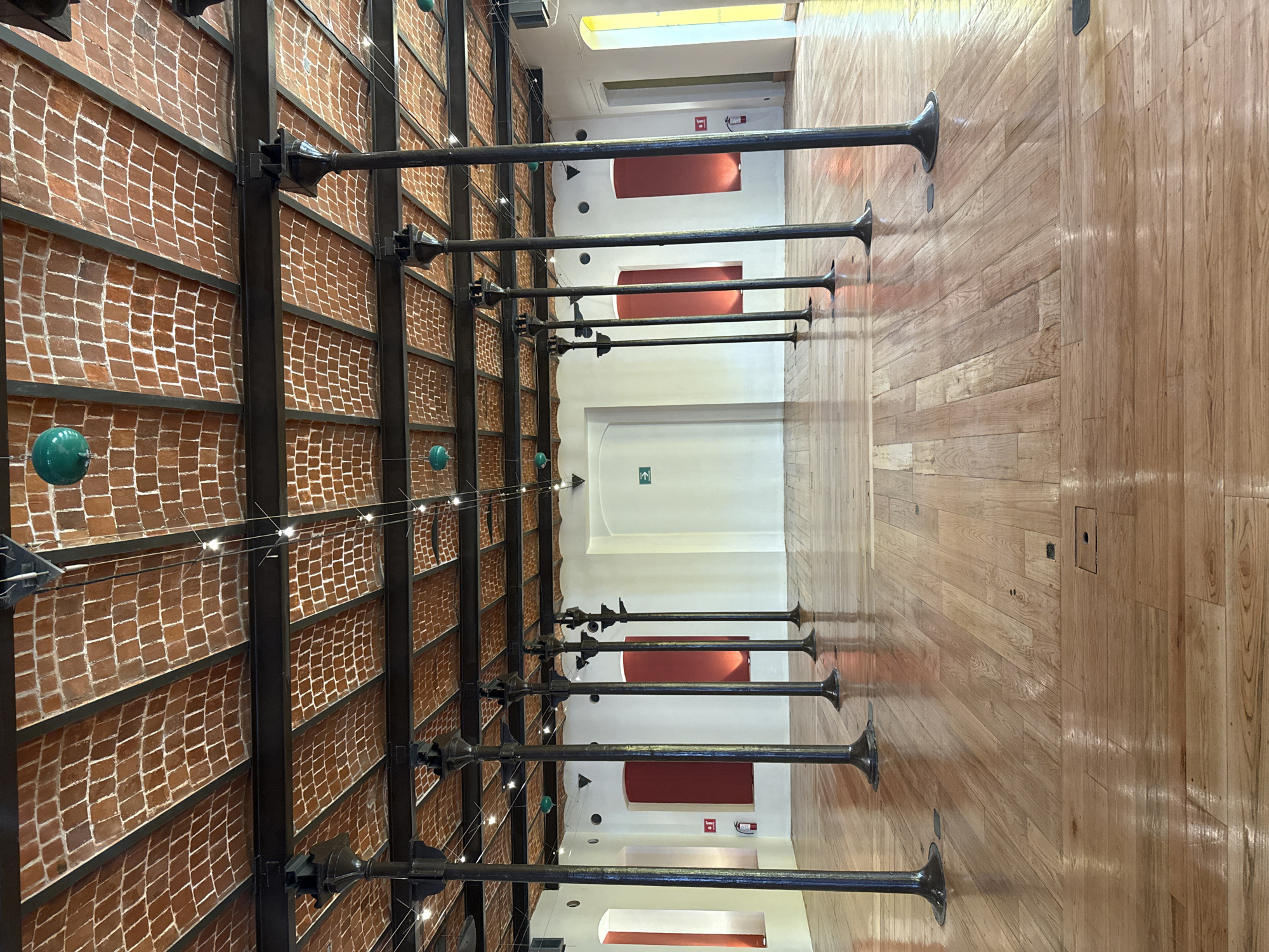 This image captures the interior of a long, high-ceilinged corridor or hall, likely within a public building or museum in Puebla, Mexico. The perspective looks down the length of the hall, which appears to be rotated 90 degrees clockwise from its natural orientation. If rotated to the correct upright position, the brick structure would be on the left wall, and the polished wooden surface would be the floor on the right.
On the left side, a prominent wall is constructed with sections of light brown bricks arranged in a distinctive diamond or basketweave pattern, framed by a grid of dark, industrial-style metal beams. Several ornate, dark metal poles extend horizontally from this brick wall into the open space of the corridor. Small, green spherical lights and delicate white string lights are suspended by wires between these metal structures.
The right side of the corridor features a highly polished, light-colored wooden floor, made of parallel planks that gleam with reflected light. A series of identical dark metal poles stand on this wooden floor, rising vertically from a flared base before extending horizontally into the corridor, mirroring the poles on the brick wall.
At the far end of the hall, a white wall is visible, punctuated by several large, recessed rectangular openings painted a deep red. Two green exit signs are mounted on this white wall; one depicts a running figure pointing left, and another points downwards. A bright yellow light fixture can be seen at the top of the ceiling. The hall is devoid of people, emphasizing its architectural design, which combines textured brick, warm wood, and robust metalwork. The ambiance is one of structured elegance and well-lit spaciousness.