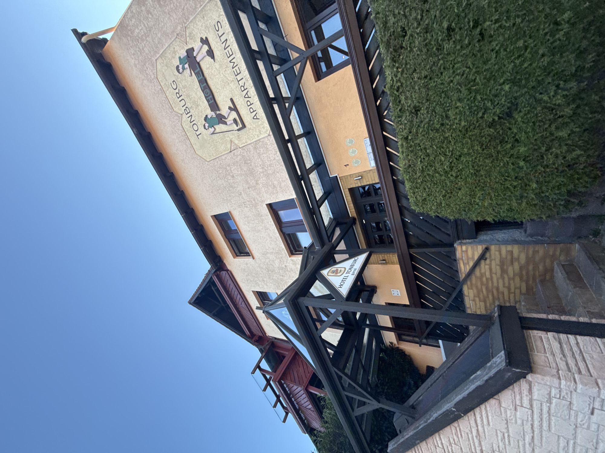 The image shows the facade of the Hotel Tonburg Appartements, located in Bad Sachsa, Germany. The building has a light-colored, textured exterior and features multiple windows. Above the entrance, a wooden structure with a glass roof partially covers a walkway. A sign for "HOTEL TONBURG garni" is visible on the structure. A prominent logo on the building's upper facade depicts two figures in profile, one holding a walking stick, with the text "TONBURG HOTEL APPARTEMENTS" around it. To the left of the entrance, a section of the building is clad in pale pink stone, and to the right, a brick wall and an overgrown green hedge are present. A set of stone stairs leads down from the entrance area. The sky above is clear and blue.