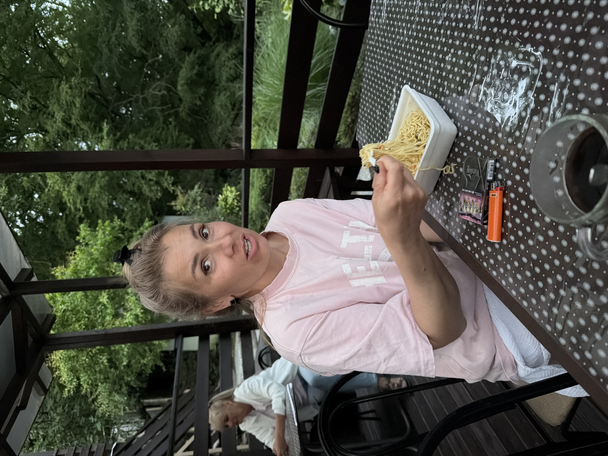 A woman with blonde hair pulled into a bun is sitting at a table with a checkered tablecloth, eating noodles from a white container with a fork. She is wearing a pink t-shirt and white shorts. To her left, an orange lighter and a pack of cigarettes are on the table. In the background, a woman in a white jacket is sitting at another table. The scene appears to be an outdoor seating area with a wooden roof and railing, surrounded by trees. The location is identified as Sochi, Russia.