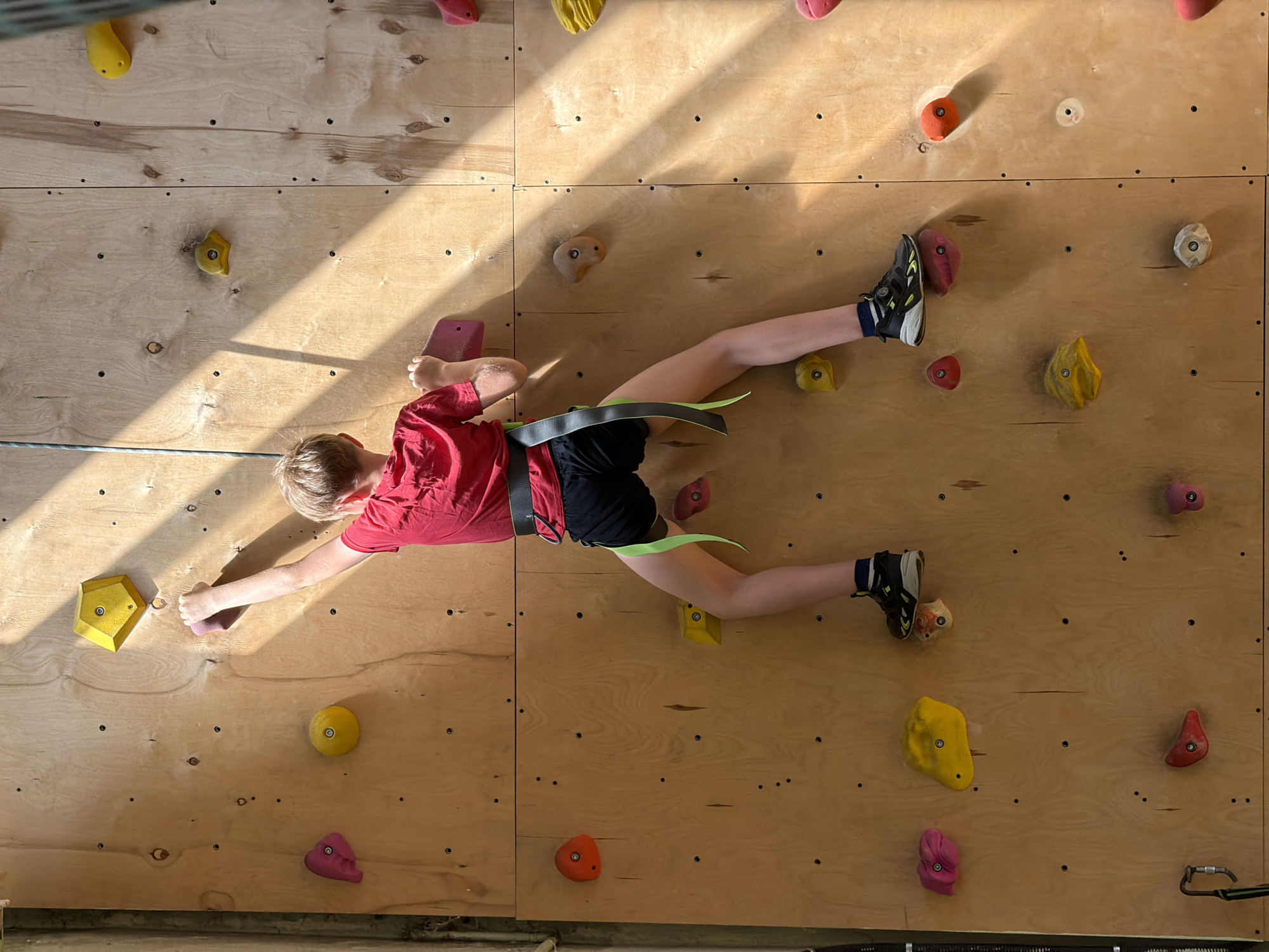 A young person in a red shirt and black shorts, wearing a climbing harness, is rock climbing indoors on a wooden climbing wall. The wall has various colored climbing holds. The location is difficult to determine without further information, but the image suggests an indoor climbing gym. There is no information indicating this picture was taken in Izhevsk, Russia.