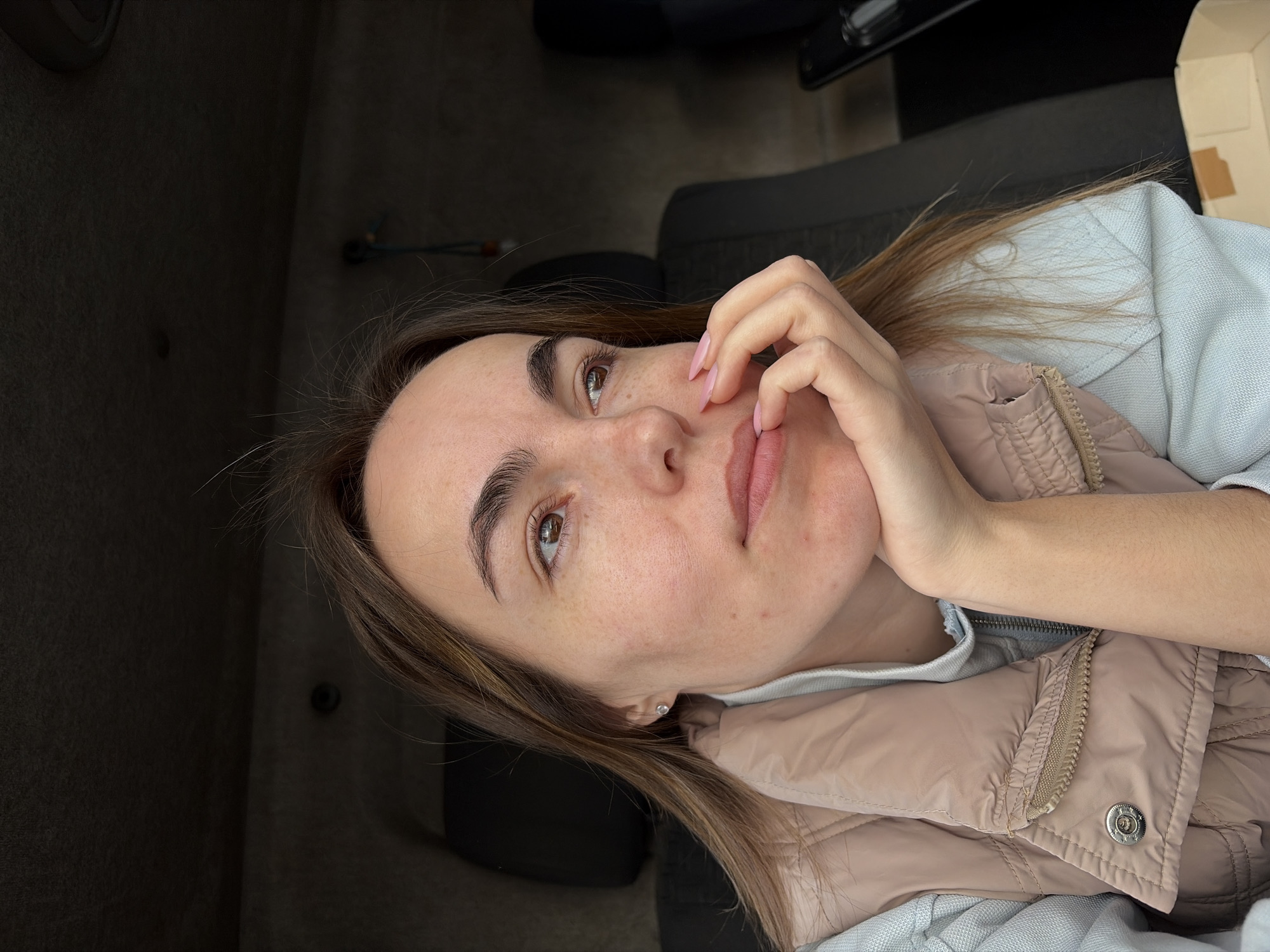 A young adult woman is shown in a close-up, tilted shot, presumably inside a vehicle. The woman has light brown hair, fair skin, and visible freckles. She wears a light blue top under a beige puffer vest. Her left hand is raised to her face, with pink nails visible. The background consists of dark grey carpeting, a portion of a black car seat, and a partially visible cardboard box in the upper right. There is no information in the image to indicate the location as Izhevsk, Russia.