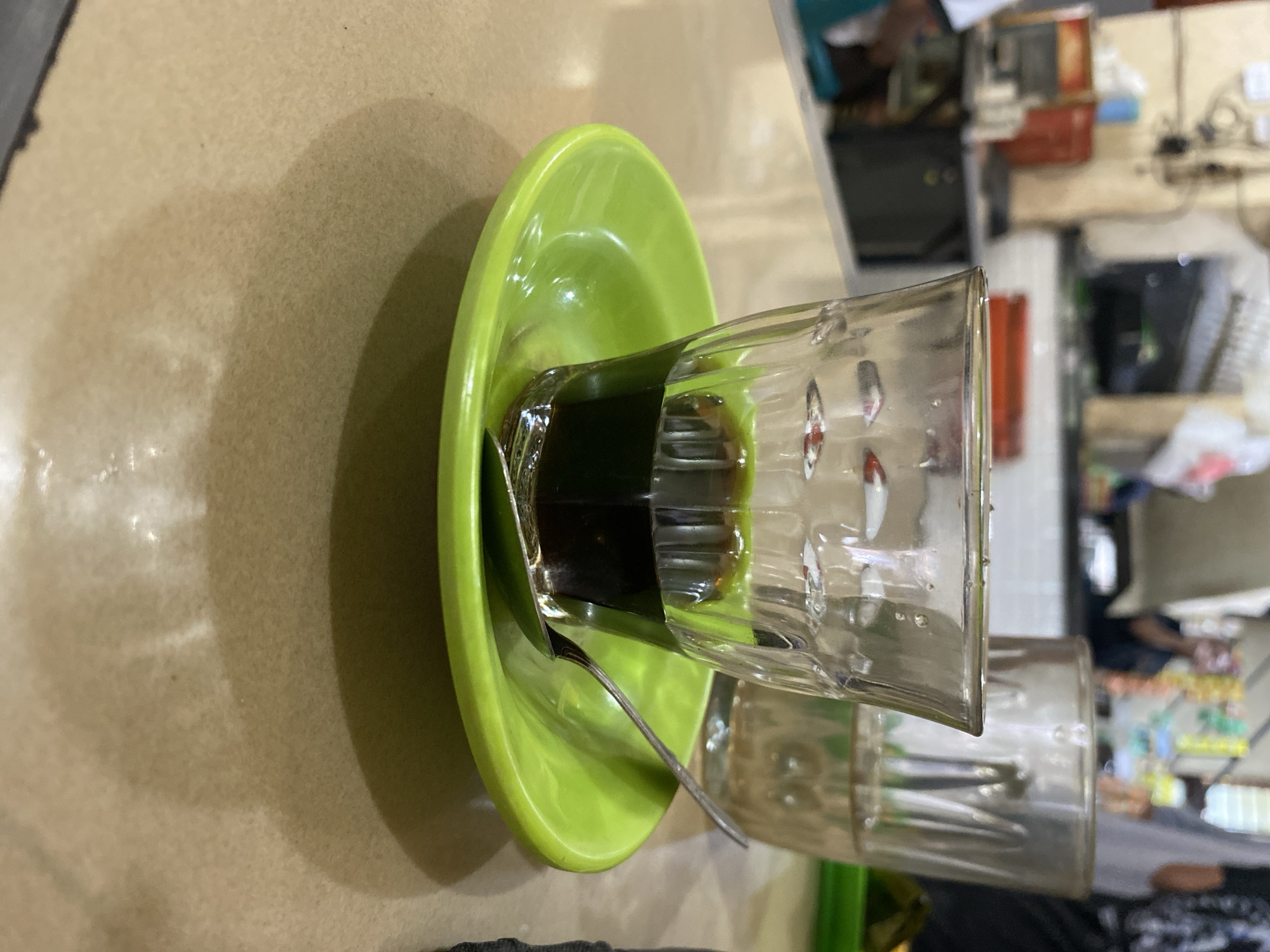 The image shows a close-up of a small glass filled with a dark liquid, possibly coffee or tea, placed on a bright green saucer. A silver spoon rests on the saucer. In the background, there is another glass with a lighter liquid, and further in the background, blurry figures of people and cafe-like setting are visible. The scene appears to be indoors, likely in a food establishment, possibly a small warung or cafe. The overall lighting is dim, with the focus on the drink and saucer. The context suggests a typical scene in a public place in Lhokseumawe, Indonesia, where such beverages are commonly served.