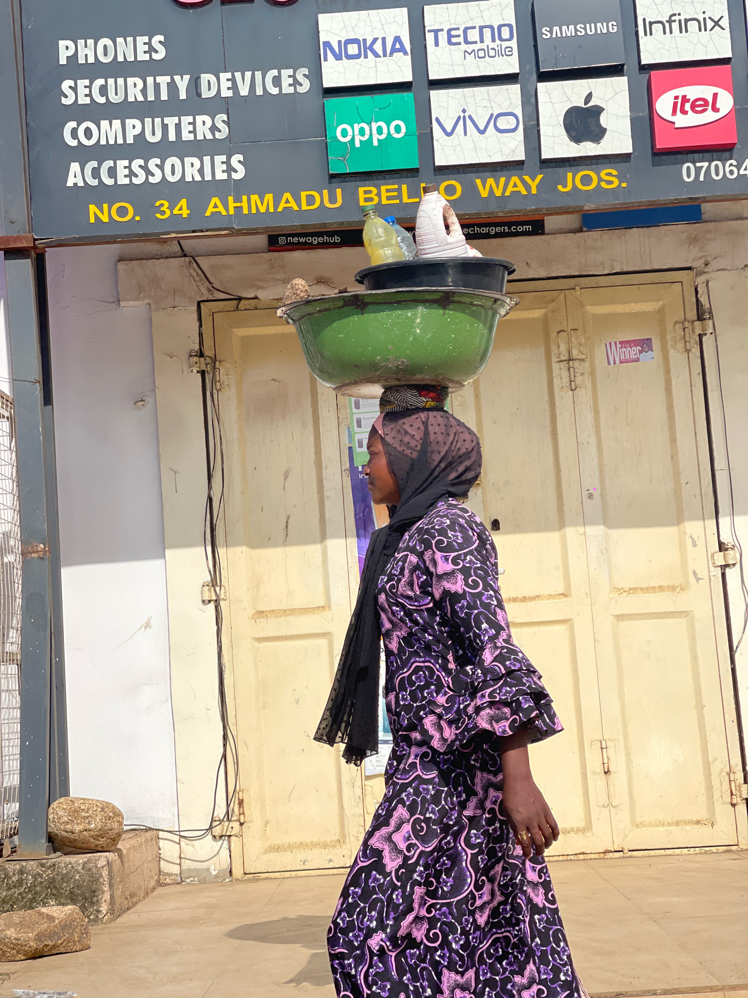 A woman in a purple and black floral dress and a black hijab balances a large green basin filled with items on her head. She is walking past a shop with a sign advertising "PHONES SECURITY DEVICES COMPUTERS ACCESSORIES" and mobile phone brands like NOKIA, TECNO, SAMSUNG, Infinix, oppo, VIVO, Apple, and itel. The shop is located at "NO. 34 AHMADU BELLO WAY JOS." The doors behind her are yellow and slightly weathered. The ground is paved with concrete. There are two rocks in the foreground.