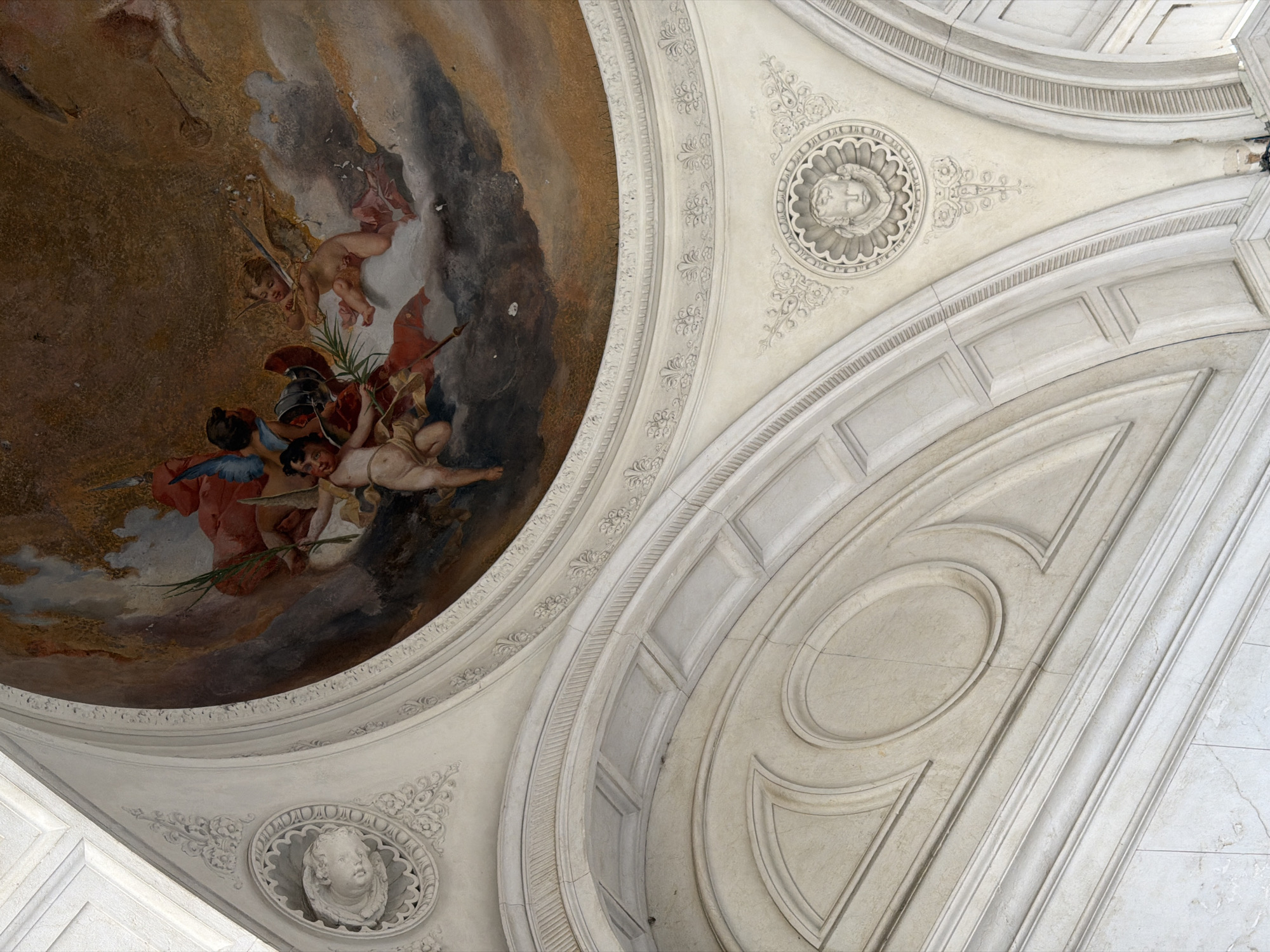 The image shows a fresco on the ceiling of a domed structure. The fresco depicts several cherubic figures in flight amidst clouds. One cherub is prominently positioned in the center, holding a branch and an arrow. Other cherubs are seen around it, some with wings and others seemingly holding weapons or banners. The fresco's colors are rich, with shades of brown, red, blue, and flesh tones dominating.
Below the fresco, the architectural elements of the dome are visible. These include plasterwork with ornate moldings and carved decorative motifs, such as rosettes and stylized floral patterns. The architecture features curved arches and recessed panels, suggesting a classical or Baroque style. The overall impression is one of grandeur and artistic embellishment.
The image's metadata indicates that the location is Mozzo, Italy. More specifically, this fresco is located within the Santuario della Madonna delle Lacrime in Mozzo, a sanctuary dedicated to the Virgin Mary.