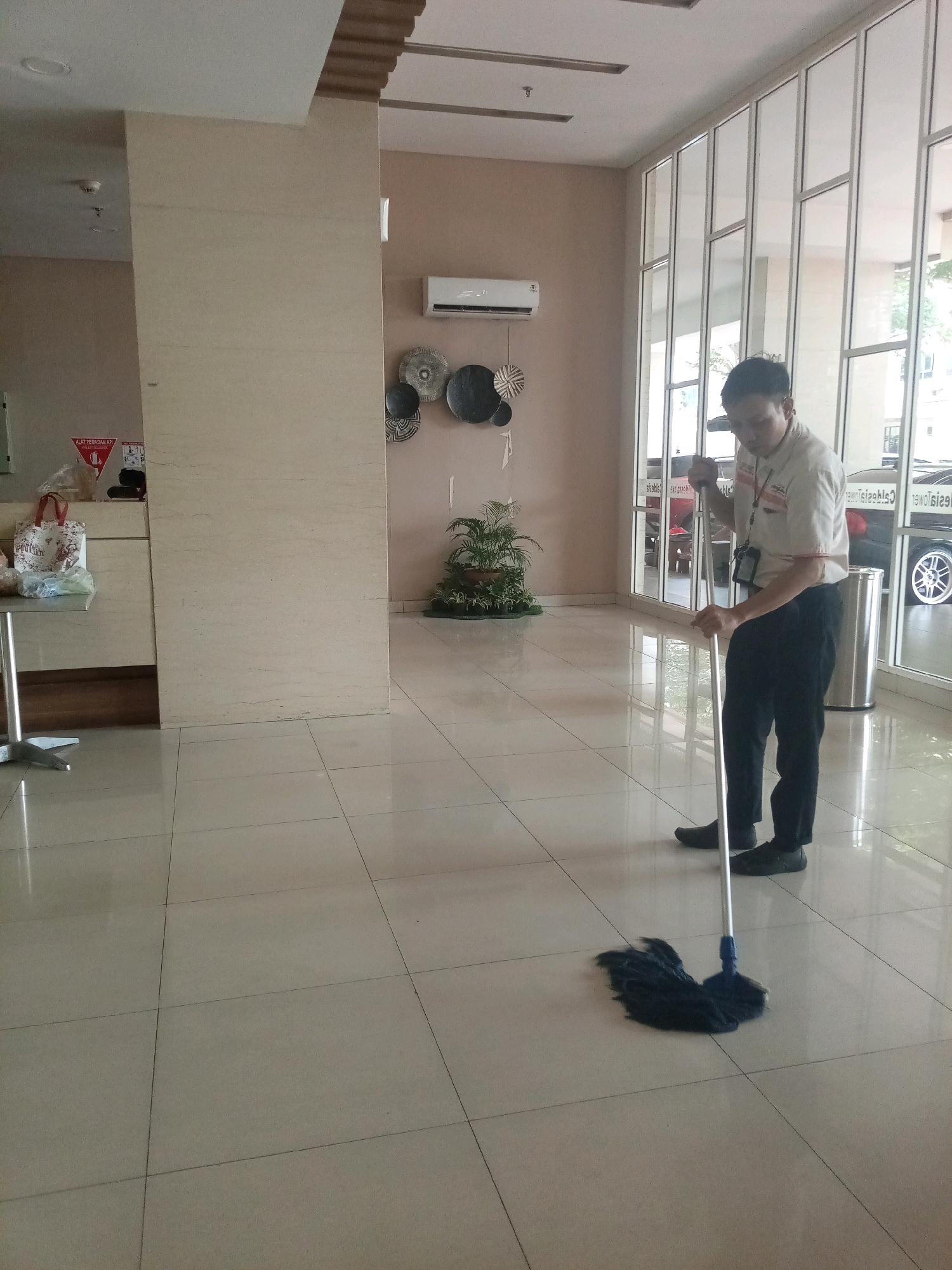 An Indonesian man, wearing a beige uniform shirt with an ID lanyard and black pants, is mopping a tiled floor in what appears to be the lobby of a building in Bekasi, Indonesia. He is holding a mop with a dark blue mop head and a silver handle. The floor is light beige and appears clean and shiny. To the right of the man, large glass windows with a silver frame look out onto a street with cars parked outside. Behind him, on the wall, are several round, decorative wall hangings. To his left, there is a large pillar and a small table with shopping bags and a fire extinguisher sign. An air conditioning unit is mounted on the wall above the decorative hangings.