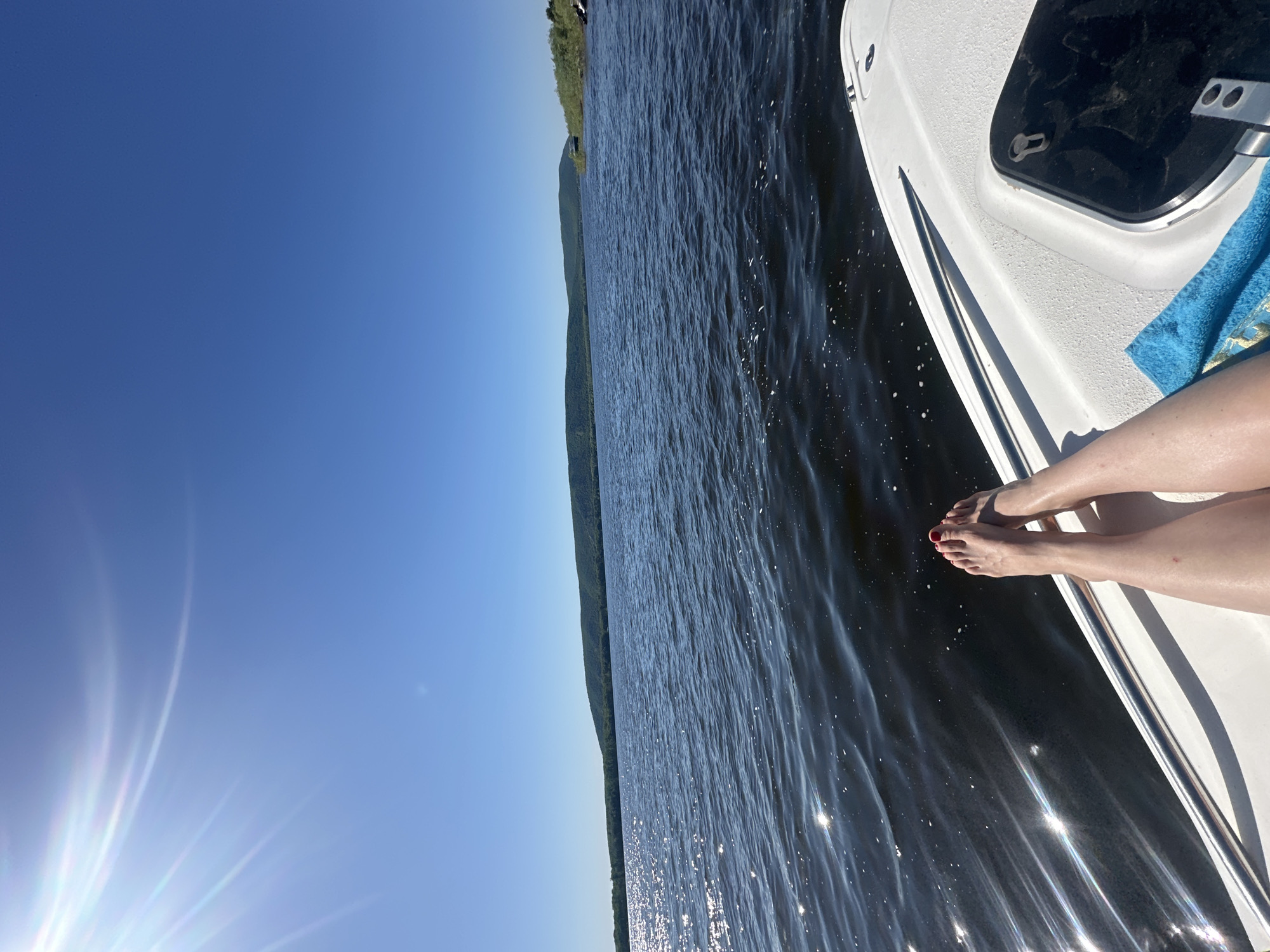 The image shows a view from a boat on a body of water. The water surface is dark and rippled, reflecting sunlight. On the right, there is part of a white boat, including a section with a dark compartment and a blue towel. Two legs with red nail polish on the toes rest on the edge of the boat. In the distance, there is a green shoreline with trees. The sky is clear and blue with a bright light source in the lower left corner. The location appears to be a natural, outdoor setting, possibly a lake or river. The city and state are unknown.