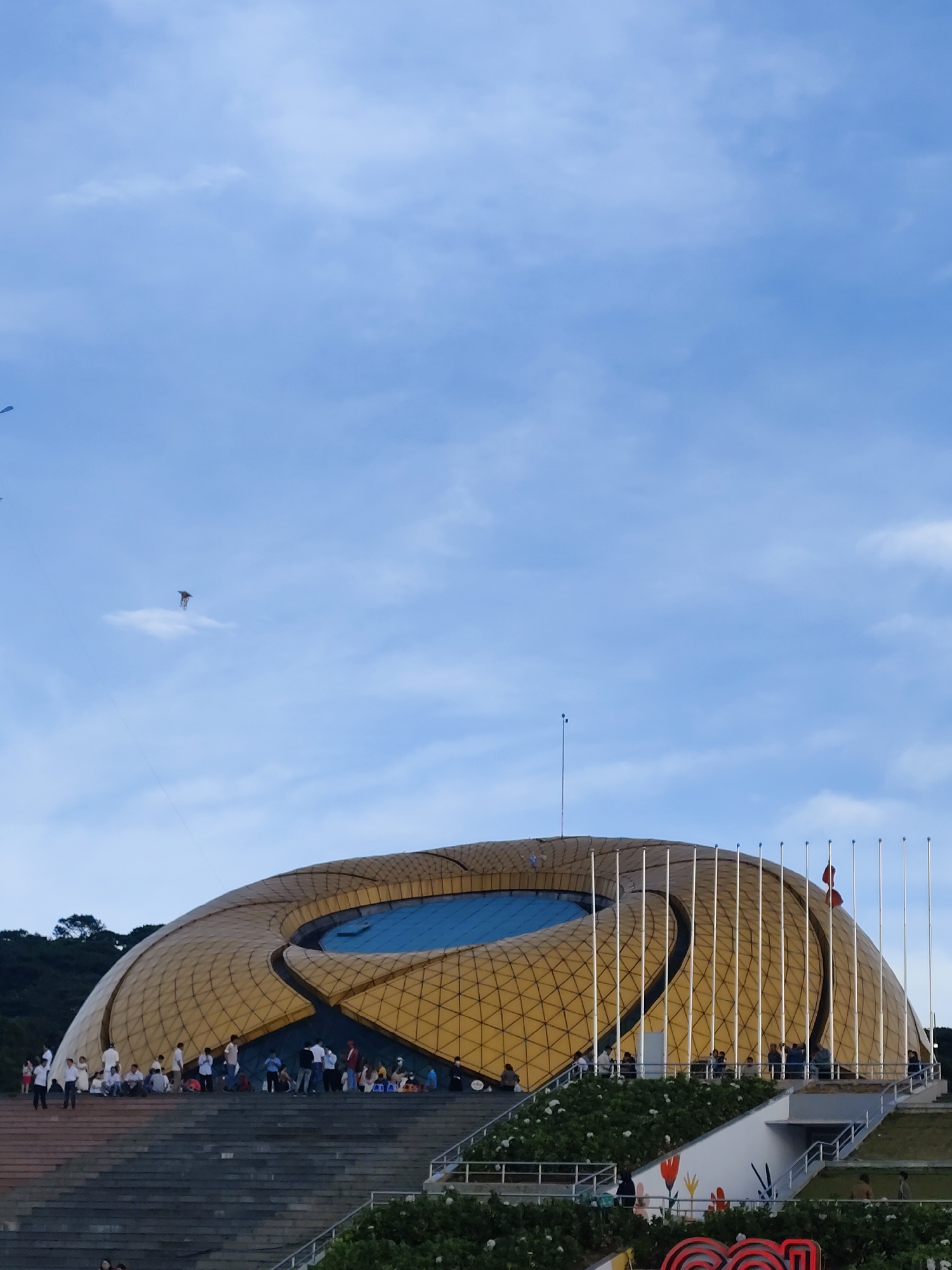 The image depicts the exterior of the Da Lat Exhibition Center, a distinctive dome-shaped building with a golden, geodesic exterior, located in Da Lat, Vietnam. The building features a large, elliptical opening with a blue pool or decorative element. A flight of wide, stone stairs leads up to the main level of the building where a crowd of people is gathered. Some individuals are sitting on the stairs, while others are standing and socializing. The sky above is a bright blue with scattered white clouds. Several flagpoles are visible on the right side of the building, with flags flying. Greenery and landscaping, including some flowering plants, surround the building. In the sky, two kites are visible, one further away and smaller, and another closer and larger, both with strings extending down.