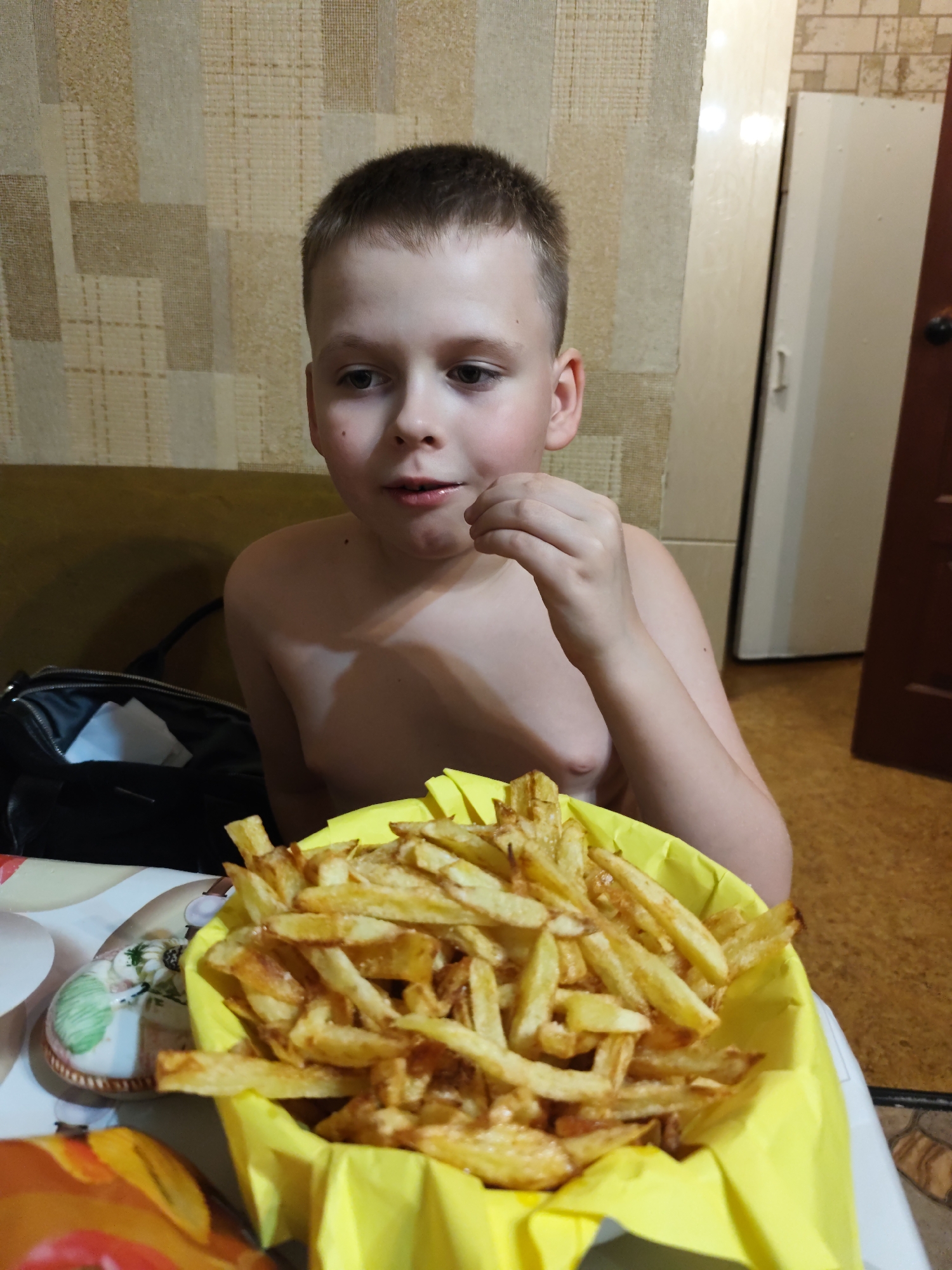 A young boy is captured in a close-up shot, looking towards the right side of the frame with a curious expression. He is shirtless, revealing his bare chest and shoulder. In front of him, a bright yellow container is overflowing with golden-brown french fries. The boy's hand is raised, with his fingers near his mouth, suggesting he might be about to eat or has just eaten a fry.
The setting appears to be indoors, possibly a dining area or kitchen, given the patterned wallpaper in the background. To the left, a black bag with some white items inside is partially visible. To the right, part of a tall, light-colored cabinet and a dark brown door can be seen. The flooring has a tiled pattern.
The lighting suggests it could be daytime or evening, but there are no strong indicators of natural light. The overall mood is casual and domestic. The image is a candid snapshot of a child and his food. There is no visible text in the image. The location context is Bor, Russia.