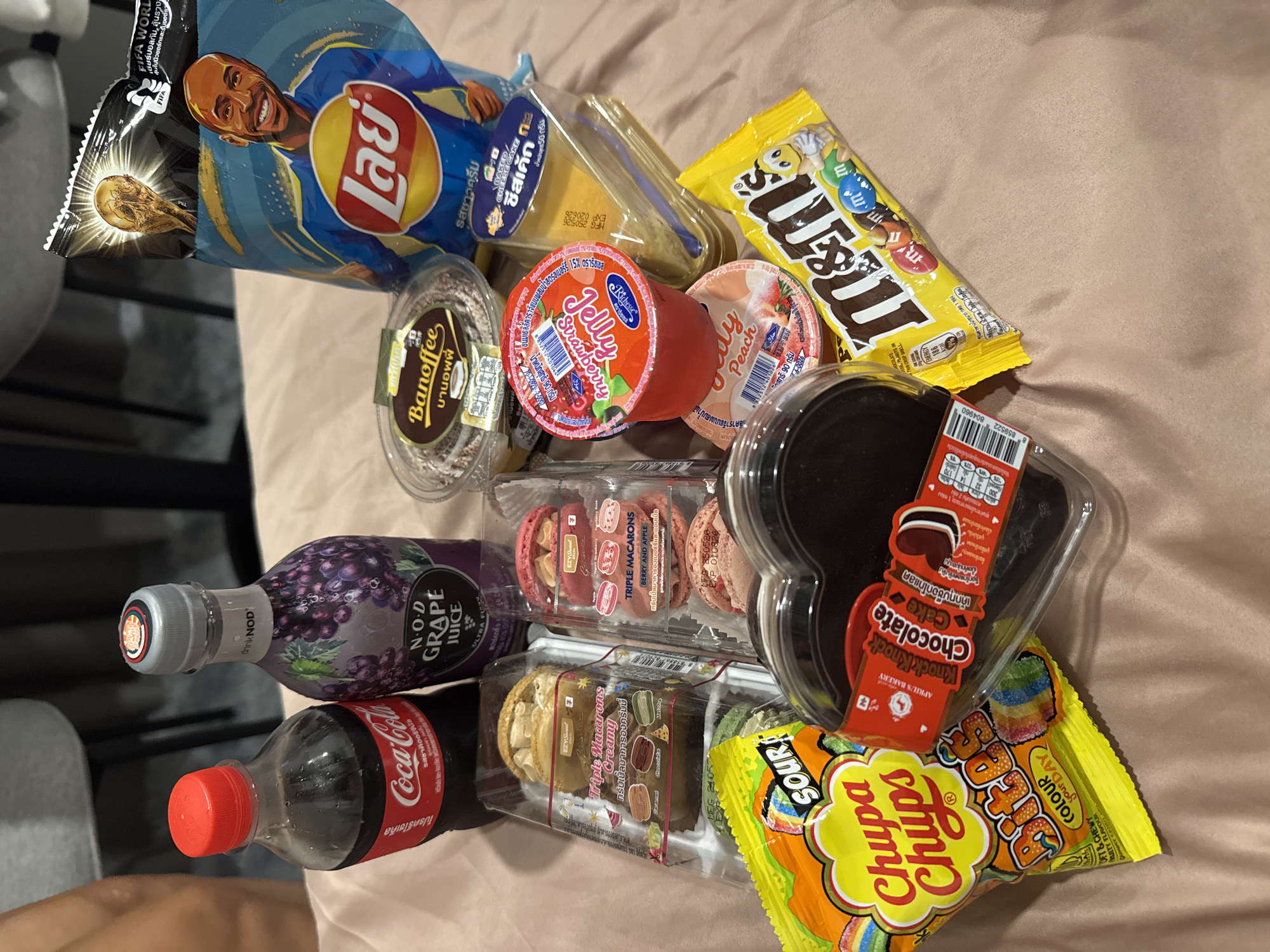 On a beige surface, a collection of snacks and drinks are arranged. The brands present include Lay's, M&M's, Chupa Chups, Coca-Cola, and Nod Grape Juice. Also visible are Thai desserts like jelly, banoffee pie, cheesecake, macarons, and heart-shaped chocolate cake. These items may have been purchased in Kathu, Thailand. The Lay's bag features an image of a soccer player holding the FIFA World Cup trophy.