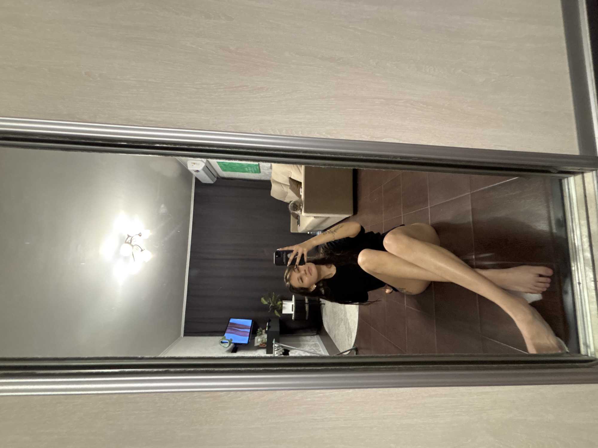 A woman with long dark hair is sitting on the floor in front of a large mirror, taking a selfie with her phone. She is wearing a black t-shirt and black shorts. Her legs are crossed and extended in front of her. The floor is tiled with dark brown tiles. In the reflection of the mirror, we can see a living room with a couch, a television, and an air conditioner. There is also a cat on the couch. The walls are a light beige color with a textured pattern. The room appears to be indoors, likely in an apartment. The location is not specified as Novokuznetsk, Russia.