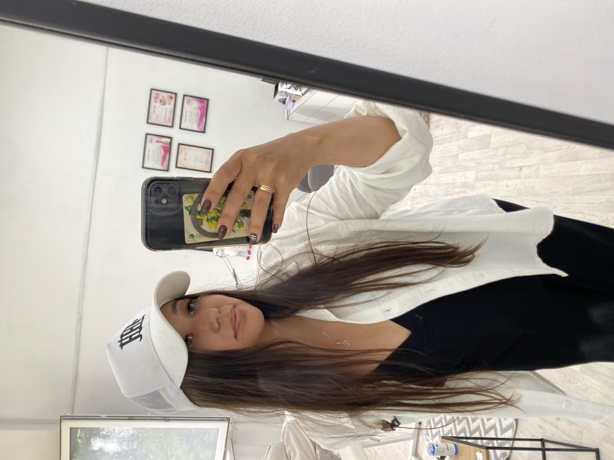 In a well-lit indoor setting, possibly a salon or studio, a woman is taking a mirror selfie. She is wearing a white trucker hat with a black logo and an open white shirt over a black tank top. Her long, dark hair is down, and she is wearing a gold necklace with a small pendant. On her right hand, she has a gold ring and dark nail polish. The mirror reflects the room behind her, which includes framed certificates on the wall and what appears to be a white cabinet. The floor is light wood laminate. The woman is positioned in the foreground, holding her black smartphone to take the picture. The overall impression is one of casual self-documentation within a professional or personal grooming space. The location is specified as Akyar, Russia, though the image itself does not provide direct visual evidence of this specific location.