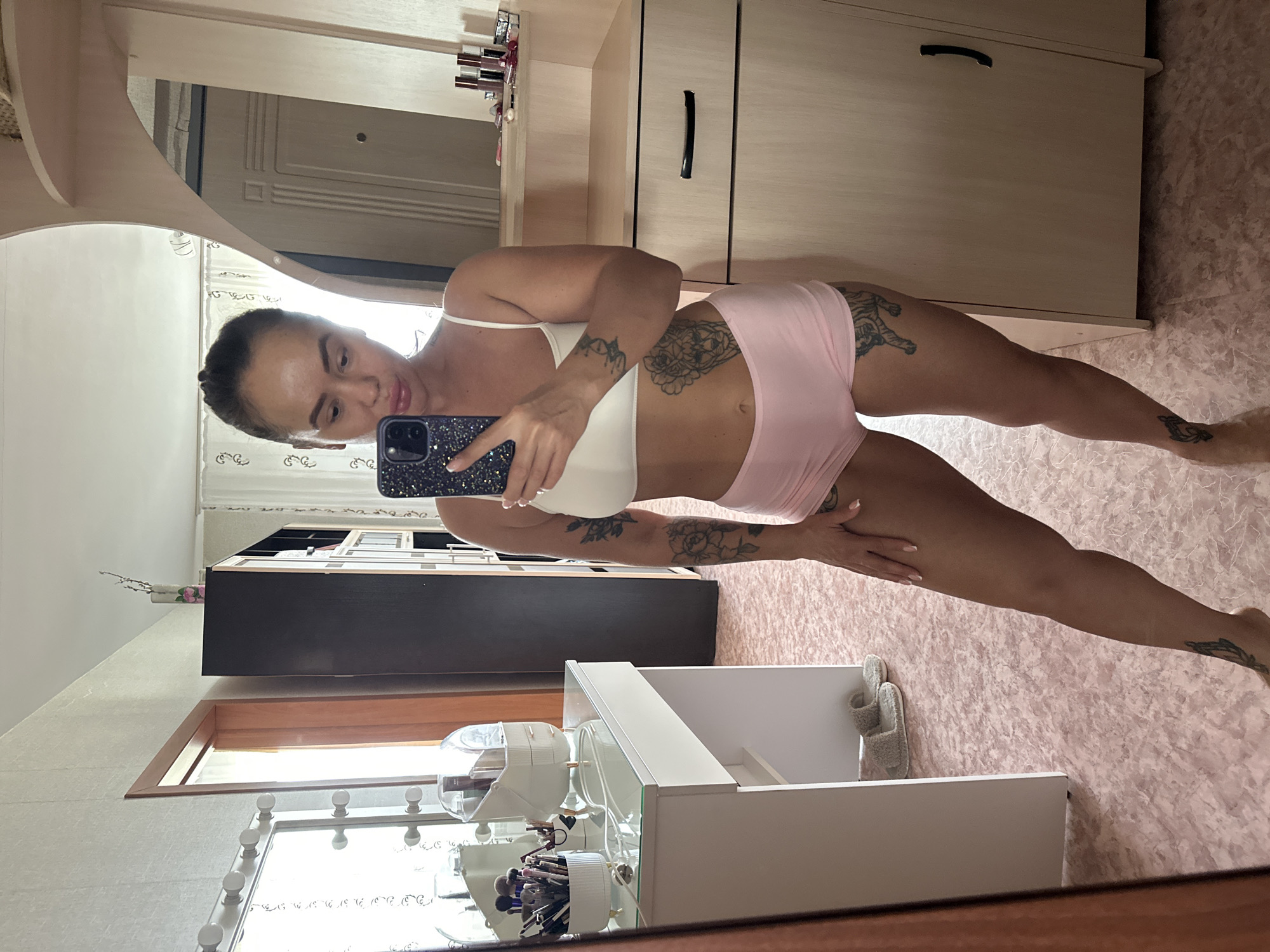 A woman is taking a mirror selfie in a room. She is wearing a white bra and pink shorts. She has tattoos on her arms and legs. She is holding a phone in her hand. In the background, there is a vanity with makeup brushes, a mirror, and a closet. The room appears to be a bedroom or dressing room. The flooring is pink with a patterned texture. The overall lighting is bright, suggesting it is daytime. The location is Yuzhnouralsk, Russia, as per the prompt.