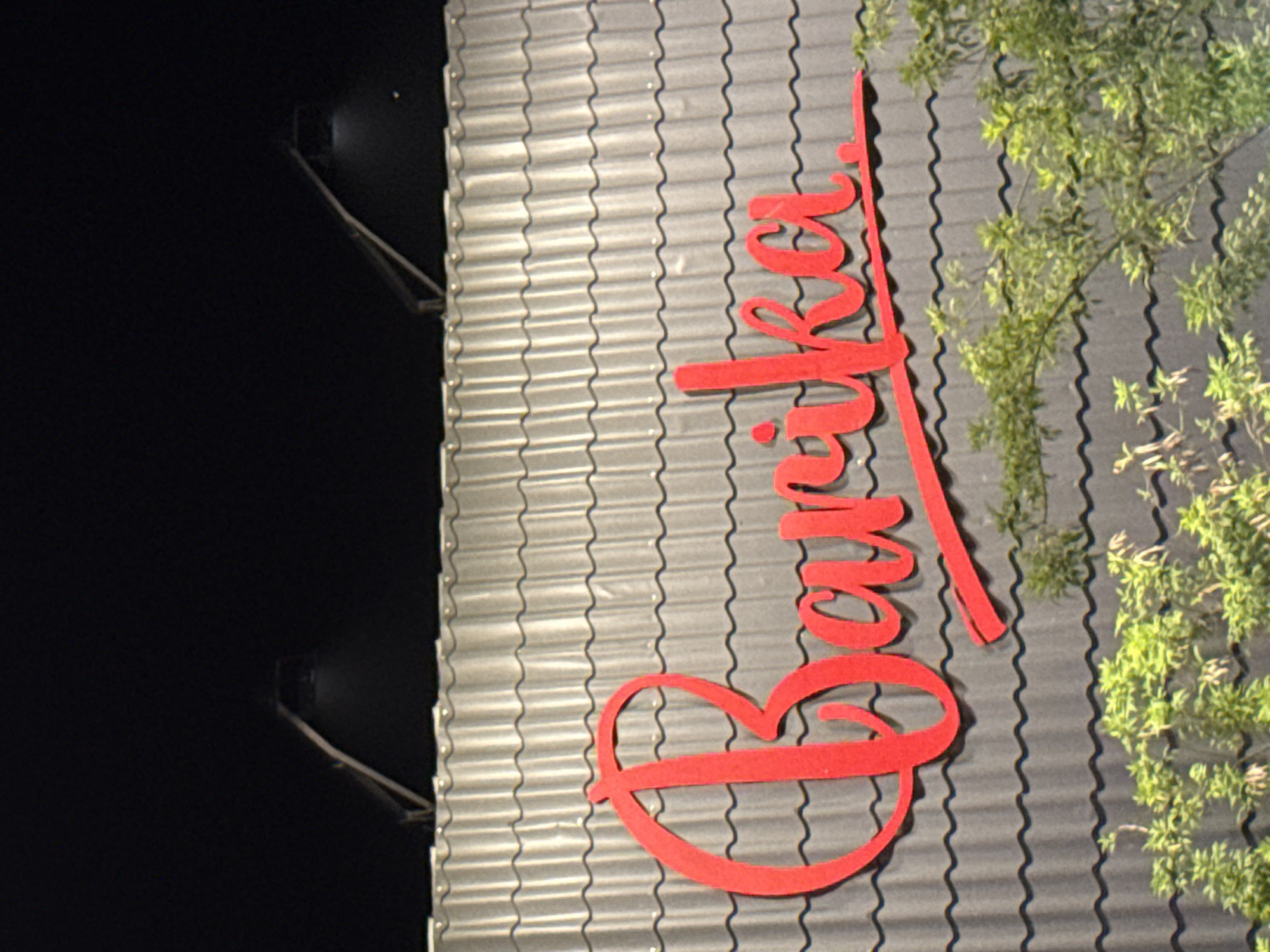 The image shows an exterior shot of a building, possibly a business, in Lambaro, Indonesia. The building has a gray corrugated metal wall with red signage spelling out "Bunker" in a cursive font. Some green foliage partially obscures the sign and wall in the upper right corner. A security light fixture is visible on the left edge of the wall against a dark background. The overall lighting suggests it is nighttime.