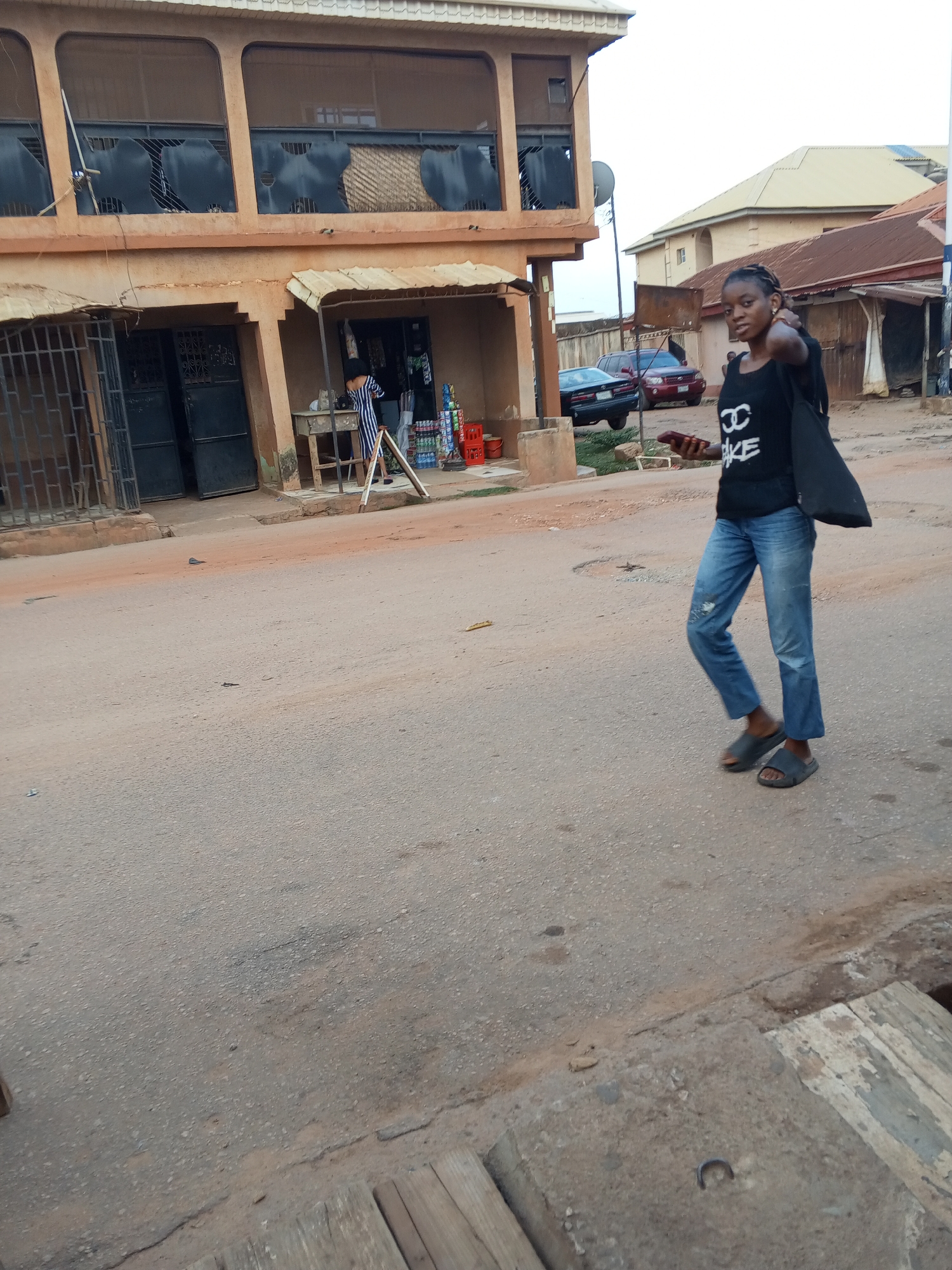 The image depicts a woman walking in a street in Sabon Tasha, Nigeria. The woman is wearing a black t-shirt, blue jeans, and sandals, and she carries a black bag. She is pointing towards the camera while holding a phone in her left hand. Behind her is a two-story building with barred windows and a small shop in front. Red crates are visible near the shop. A couple of cars are parked further down the street. The street appears to be made of asphalt and is somewhat worn. Other buildings are visible in the background.