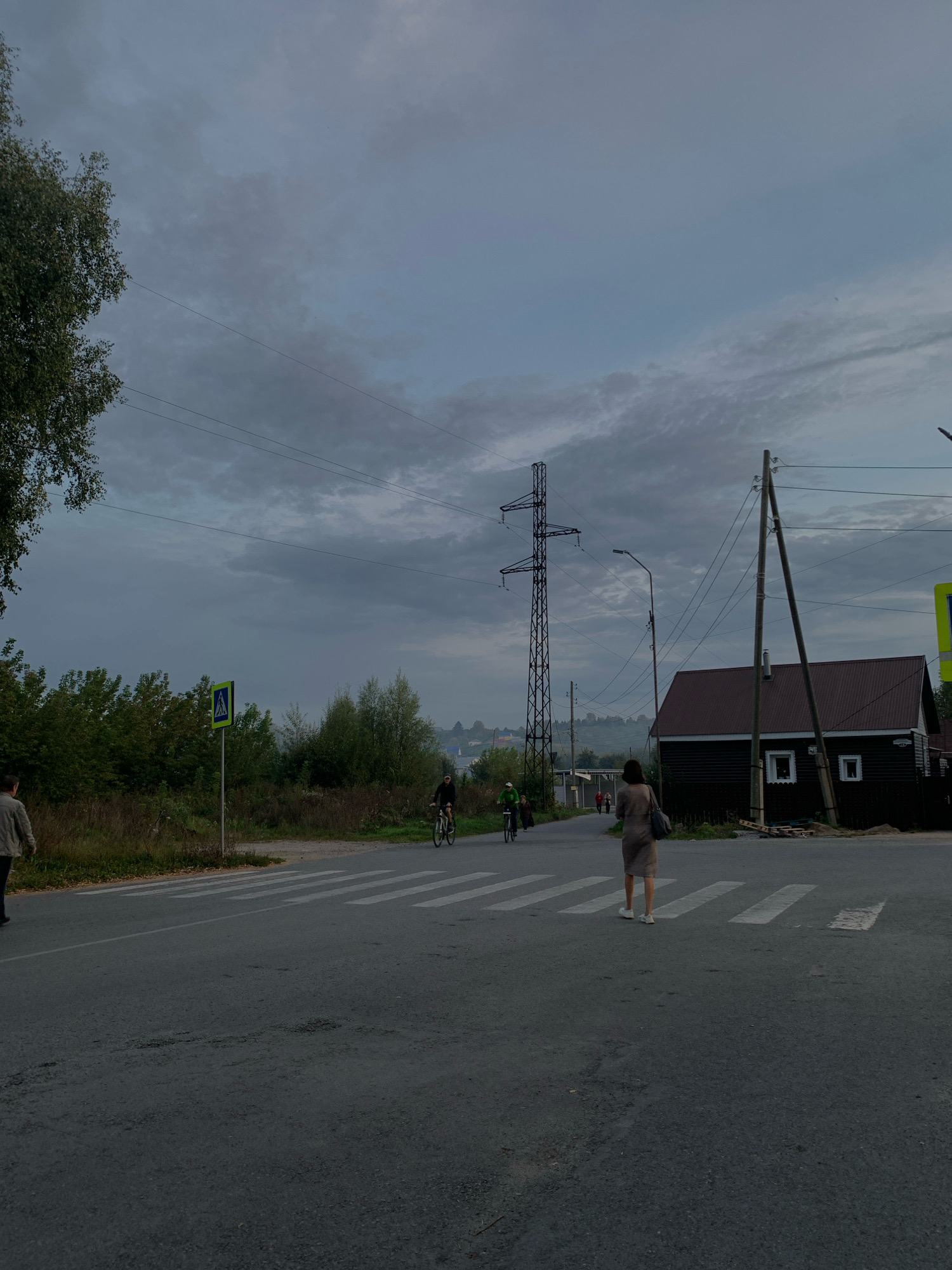 The image depicts a street scene in Asha, Russia, under an overcast sky. In the foreground, a pedestrian crossing is visible on a paved road. A woman wearing a light-colored dress and carrying a dark bag is walking across the pedestrian crossing from right to left. To the left of the crossing, a man in a light-colored jacket is walking away from the camera. Further in the background, on the right side of the road, a dark-colored wooden house with a brown roof and white-framed windows stands. Two wooden utility poles with numerous electrical wires are prominent in the scene. A tall, lattice metal electrical tower is located to the left of the house. Several people are seen in the distance on bicycles or walking, and a yellow pedestrian crossing sign is posted on the left side of the road. Lush green trees and vegetation line the sides of the road, and distant hills can be seen under the cloudy sky.