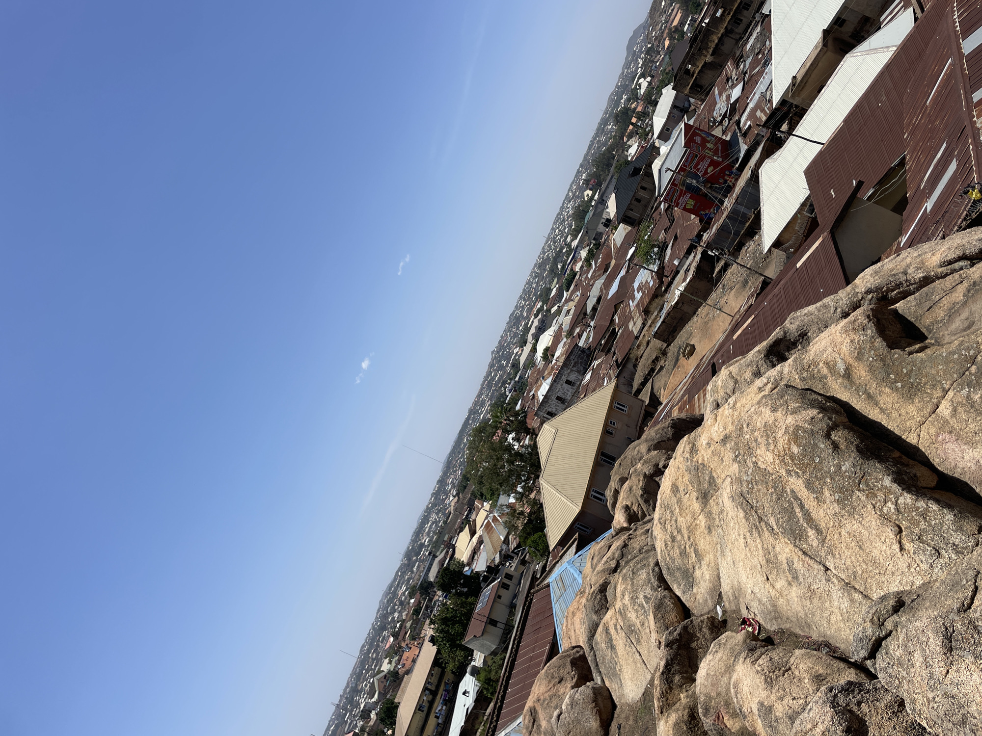 The image captures an elevated view of a densely populated area in Jos, Nigeria, taken from a rocky outcrop. The foreground features large, weathered boulders with visible crevices and textures. Beyond the rocks, a sprawling cityscape unfolds, characterized by tightly packed buildings with corrugated metal roofs, varying in color from brown and rust to white and blue. A few taller structures are interspersed among the predominantly low-rise buildings. In the distance, the city blends into the horizon, with faint outlines of hills or higher terrain. The sky is a clear, light blue, with minimal cloud cover. The photograph appears to be taken during daylight hours, evidenced by the bright sunlight casting shadows across the scene. The angle of the photograph gives a sense of looking down upon the city from a considerable height.