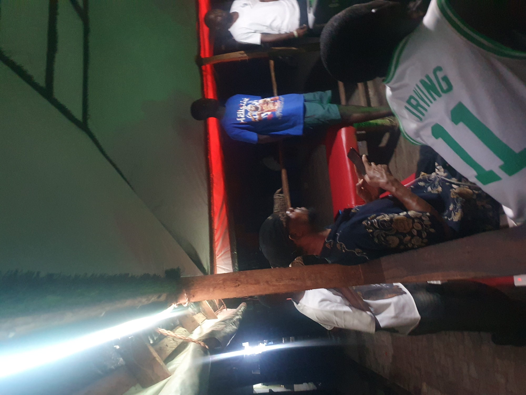 Several people are gathered on a wooden platform, with a structure possibly providing cover above. One individual wears a white jersey with "IRVING" and the number "11" in green. Another person wears a blue t-shirt with a graphic. The location is inferred to be Lokoja, Nigeria. The surrounding environment appears dimly lit. Some people are sitting, and one is using a cell phone.