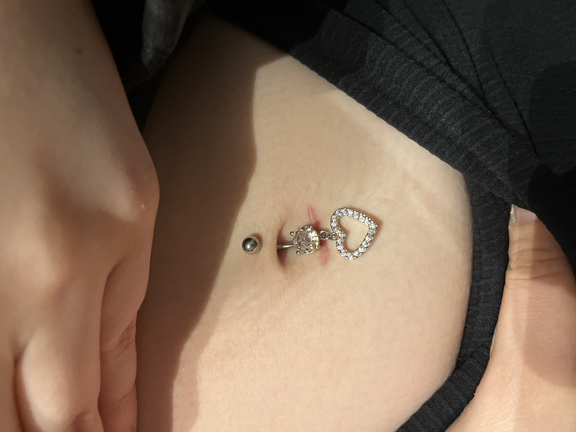 This close-up, high-angle image captures a person's torso, specifically focusing on their navel and a belly button piercing. The subject has a light skin tone. A prominent silver-toned navel ring is centered in the belly button, featuring a large, clear, round faceted gemstone at the bottom and a smaller, plain silver ball at the top. Attached to the bottom gemstone is a dangling heart-shaped charm, intricately encrusted with numerous small, clear, sparkling gemstones.
Immediately above the top silver ball of the piercing, a noticeable, slightly raised and reddish mark or scar is visible on the skin, suggesting a recent piercing, a healing process, or irritation. The surrounding skin appears smooth.
Portions of the person's hands are visible on either side of the frame. On the left, several fingers and part of a palm rest gently on the abdomen. On the right, fingertips are seen, possibly holding or adjusting black fabric, which is also visible along the top and right edges of the frame, likely being a part of clothing such as a top or bra.
The image provides an intimate view of the body modification, likely taken indoors in Chaikovsky, Russia. There is no visible text.