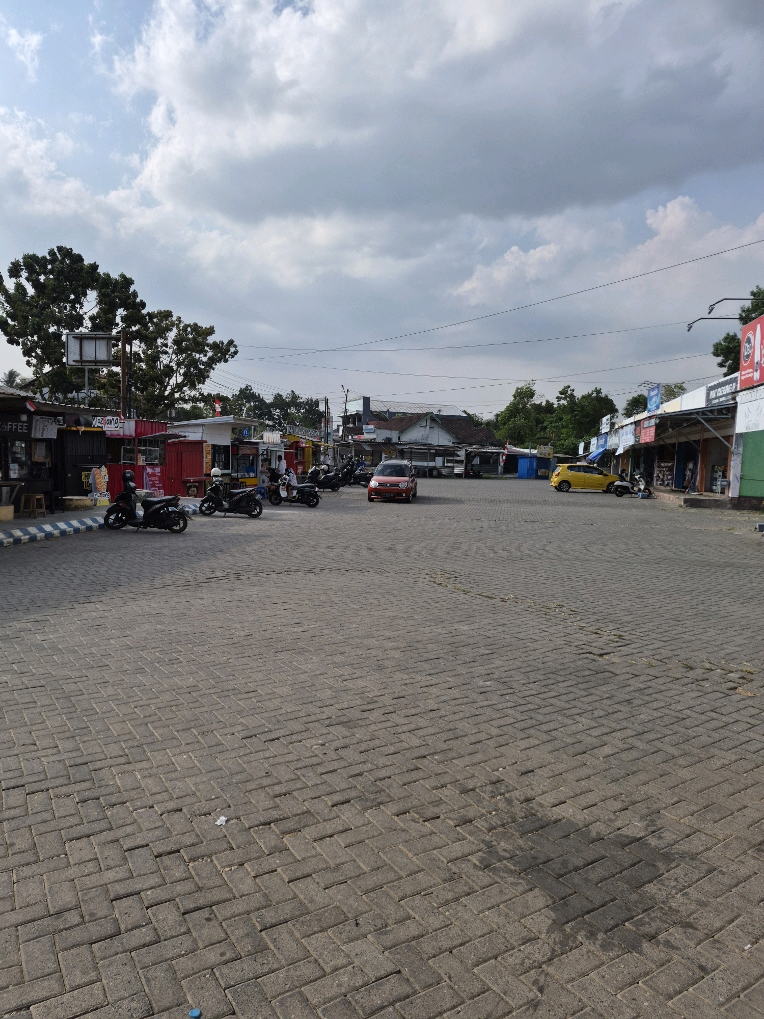 The image depicts an outdoor scene in Banjarbaru, Indonesia, likely a commercial area or parking lot. The foreground is dominated by a paved surface made of interlocking gray rectangular bricks, laid in a herringbone pattern. Scattered across this pavement are a few dark stains, possibly from oil or water, and a small piece of white debris.
In the middle ground, several small shops or food stalls are visible, constructed from brightly colored metal containers, predominantly red and white. These stalls have awnings and are lined up along a paved area. In front of these stalls, several motorcycles are parked, some with riders. One red car is parked on the left side of the image, and a yellow car is parked further back on the right.
In the background, more buildings and trees can be seen. The sky is partly cloudy, with large gray and white clouds against a light blue sky. Overhead, power lines are strung across the scene. The overall impression is a bustling commercial or roadside stop area in an Indonesian city.