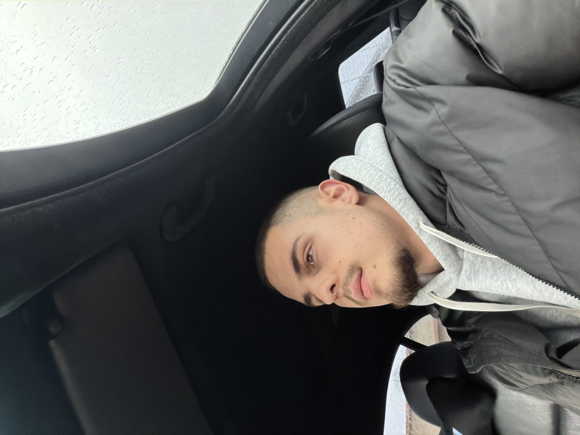 A man sits inside a car, likely in Plovdiv, Bulgaria, given the request. He is wearing a gray hoodie under a black puffer jacket. The car has a sunroof, visible with raindrops on the glass. The man is looking towards the camera. The interior of the car is dark, with the sun visor visible in the lower left.