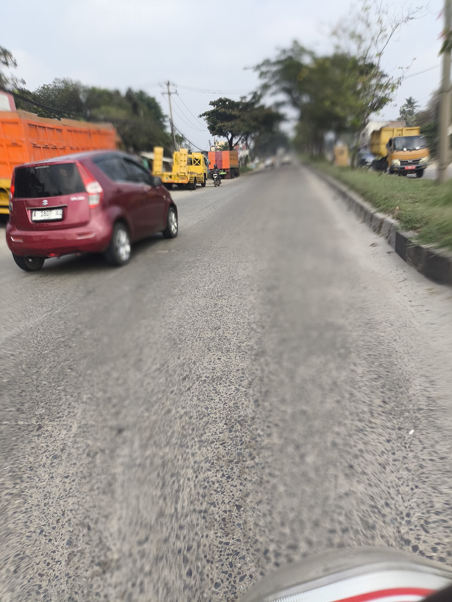 The image depicts a street scene in Cilegon, Indonesia, characterized by a road with asphalt paving. The scene is dominated by vehicles, including a red car in the foreground with license plate "A 1807 LL," and several trucks in the background. One truck is bright orange, another is yellow, and a smaller yellow truck with a cage is also visible. There is also a motorcycle with a rider. The environment includes grassy verges along the side of the road, trees, and utility poles. The sky is overcast. The image appears to have been taken from a moving vehicle, as indicated by the motion blur in the foreground.