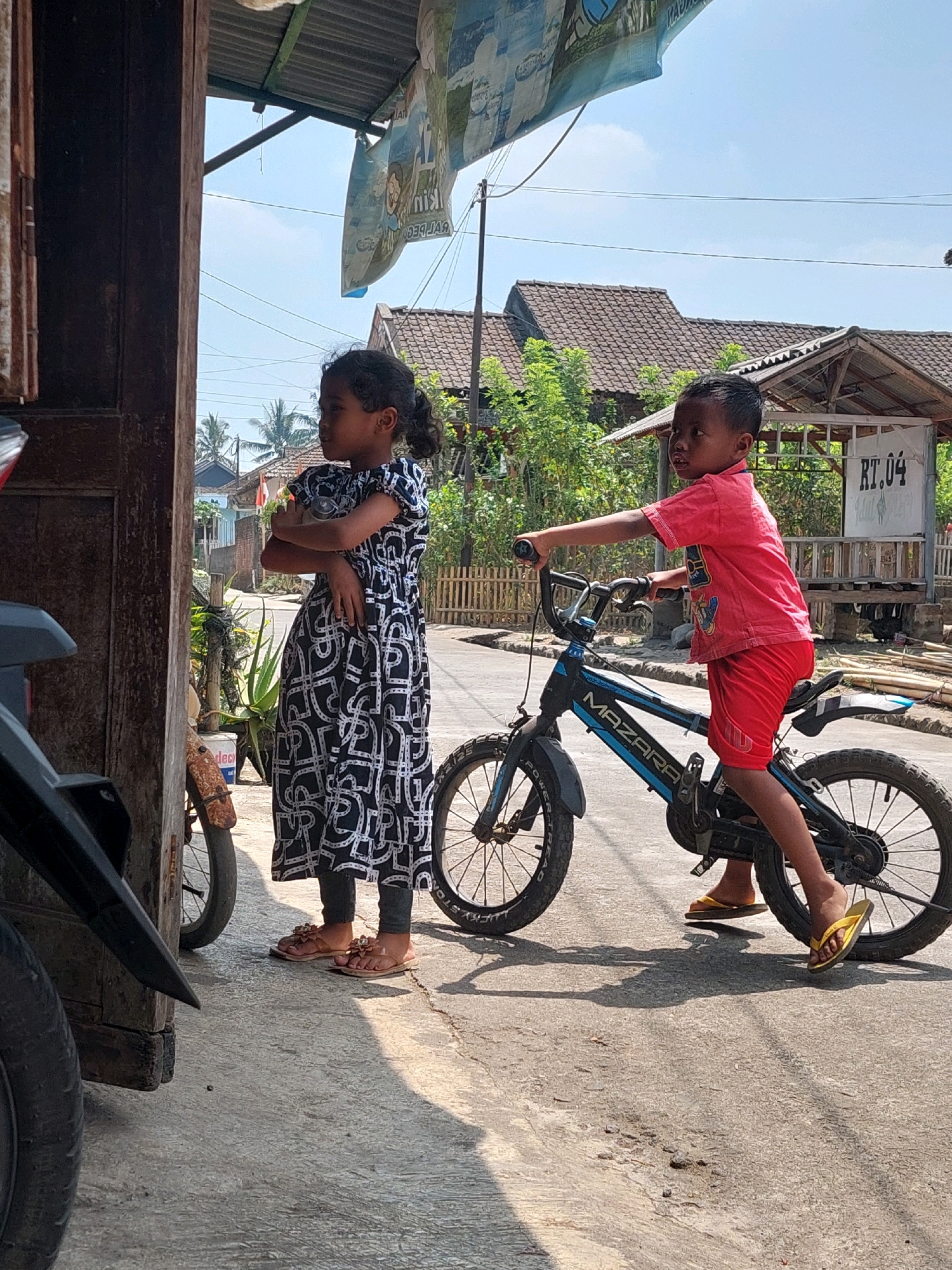 In Jogonegoro, Indonesia, on a sunny day, two children are outdoors. A young girl, wearing a patterned dress and sandals, stands with her arms crossed. Beside her, a boy is seated on a blue and black bicycle, dressed in a red shirt and shorts, and yellow flip-flops. He is holding the handlebars. The background shows houses with tiled roofs, lush green trees, and a street. Part of a motorcycle is visible to the left of the frame.