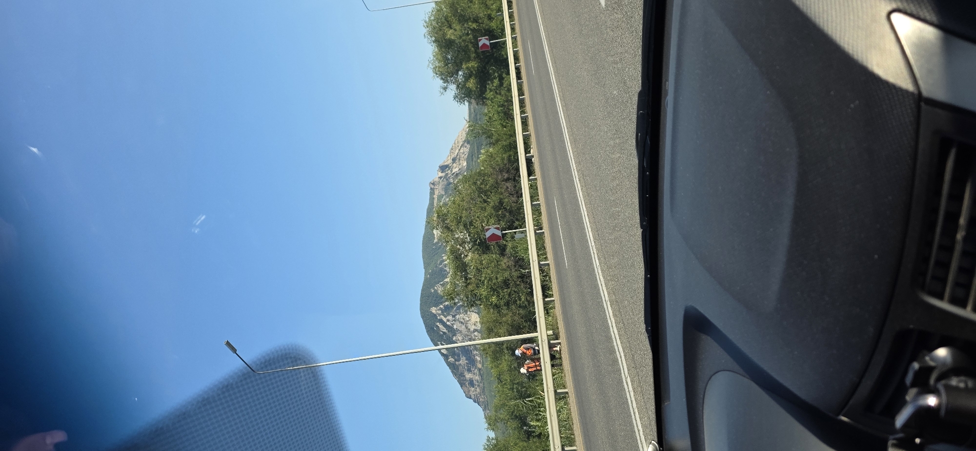 A wide-angle, low-angle, outdoor, daytime, eye-level shot captures a road in the vicinity of Zmeyka, Russia. The image features a mountain range in the background, with lush green trees lining the roadside. Two individuals, wearing orange vests and backpacks, are seen walking along the shoulder of the road. A street lamp stands tall on the left side of the image, and two traffic signs indicating a curve to the right are visible. The foreground is dominated by the dashboard of a vehicle, suggesting the photo was taken from inside a car. The sky is a clear, bright blue.