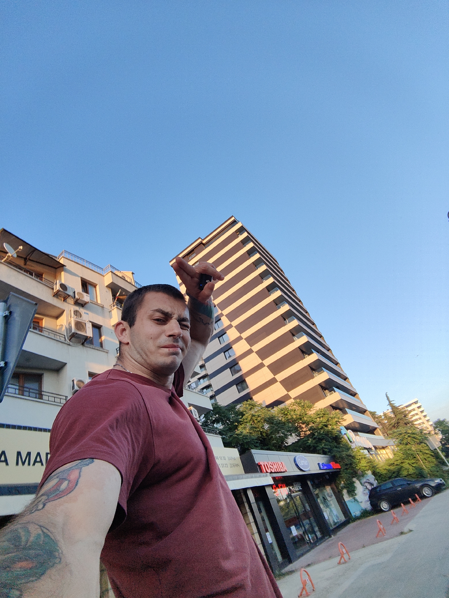 A low-angle, wide shot shows a man standing in front of buildings in Varna, Bulgaria. The man, wearing a maroon t-shirt and with tattoos on his arm, is positioned in the foreground on the left. He is looking towards the camera with a slightly distorted facial expression and has his right hand raised. Behind him, to the left, is a multi-story building with balconies and air conditioning units. To the right of this building, and further back, is a tall, modern high-rise with a distinctive striped facade of brown and beige. In front of the high-rise, at street level, are shops with visible signage, including "TOSHIBA" and "Cartier". A black car is parked on the street, and orange traffic cones are placed along the sidewalk. The sky is clear and blue, suggesting it is daytime. The overall scene depicts an urban street environment.