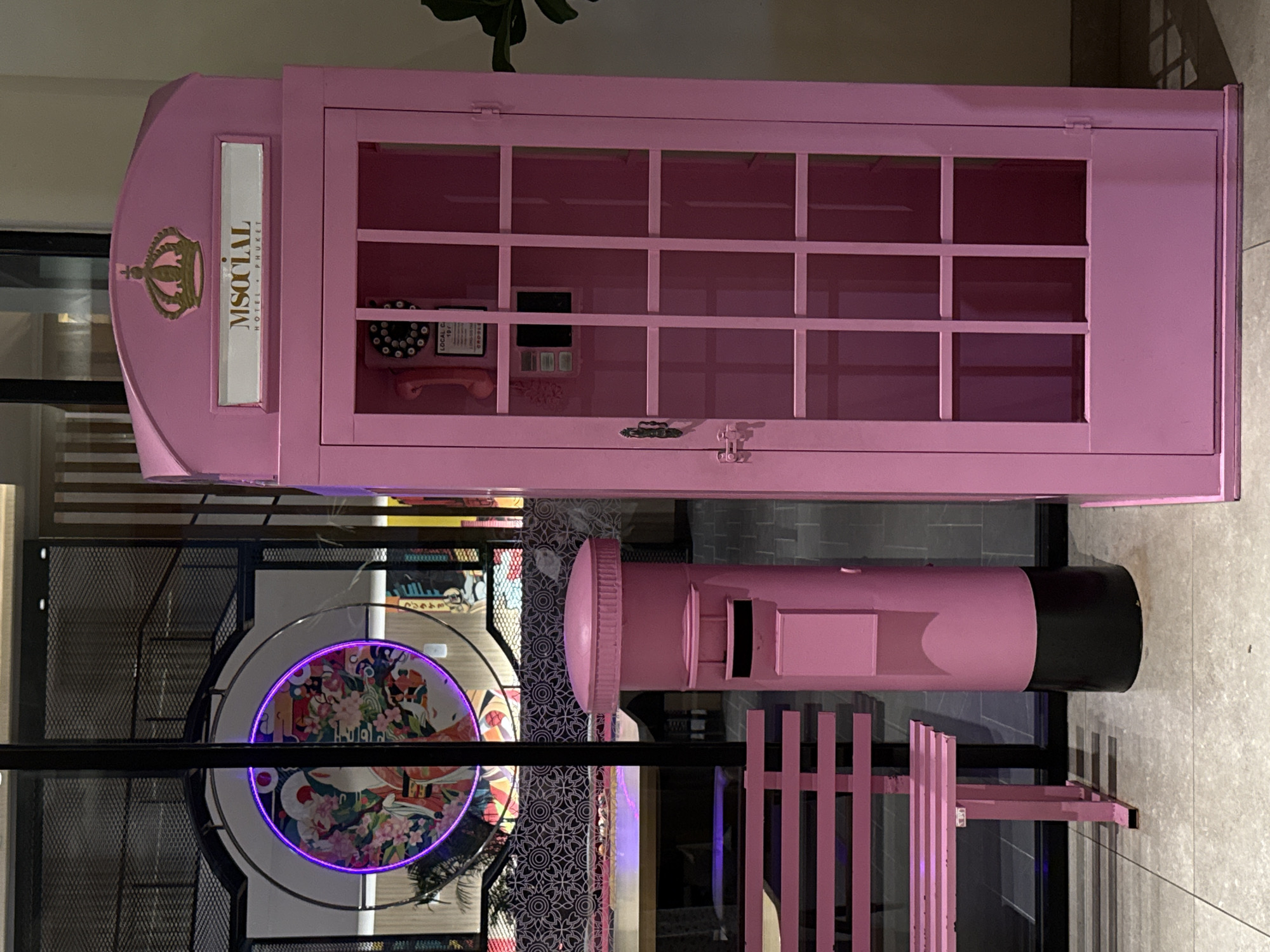 The scene features a pink M Social Hotel Phuket telephone booth with a vintage phone and grid window design. Adjacent to it is a matching pink postbox with a black top. Below, there's a circular lighted art piece. The setting appears to be inside a hotel or lobby area in Patong, Thailand.
