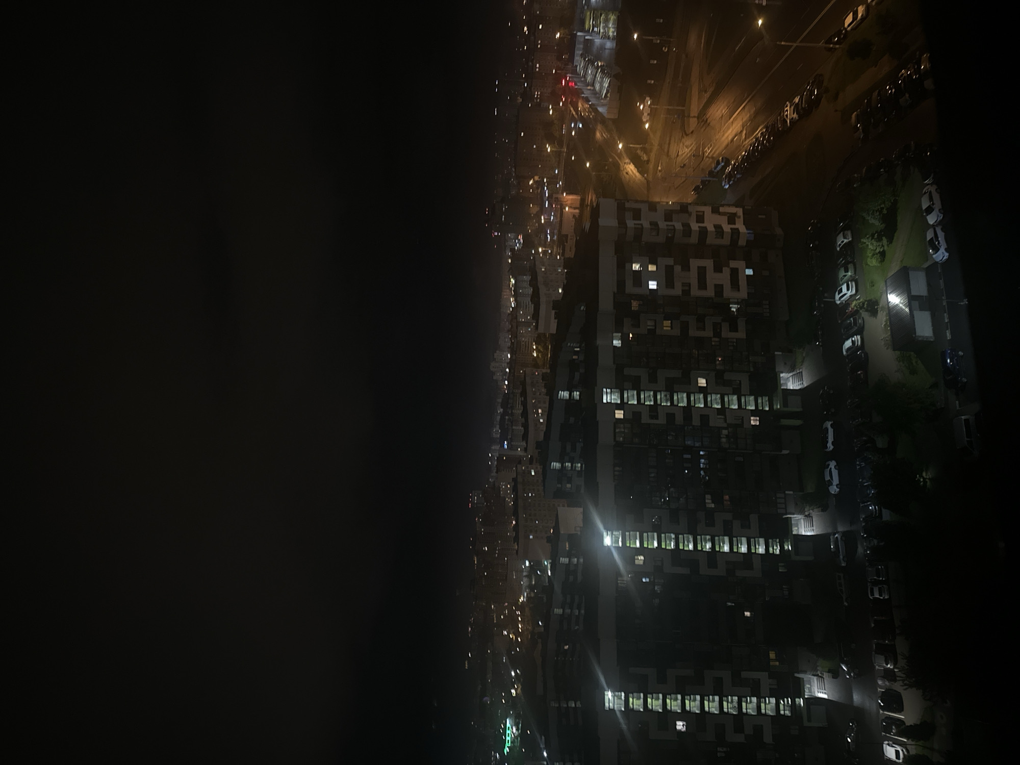 An aerial view looking down onto a cityscape at night. A tall apartment building with many illuminated windows stands prominently in the center-right. To the right of the building is a street with parked cars. Above the apartment building, there is another street illuminated by street lights. Across the top is a series of lights. To the left, the image is dominated by darkness. The location is likely in Бараўлянскі сельскі Савет, Belarus, given the prompt. The scene depicts a modern urban environment at night.