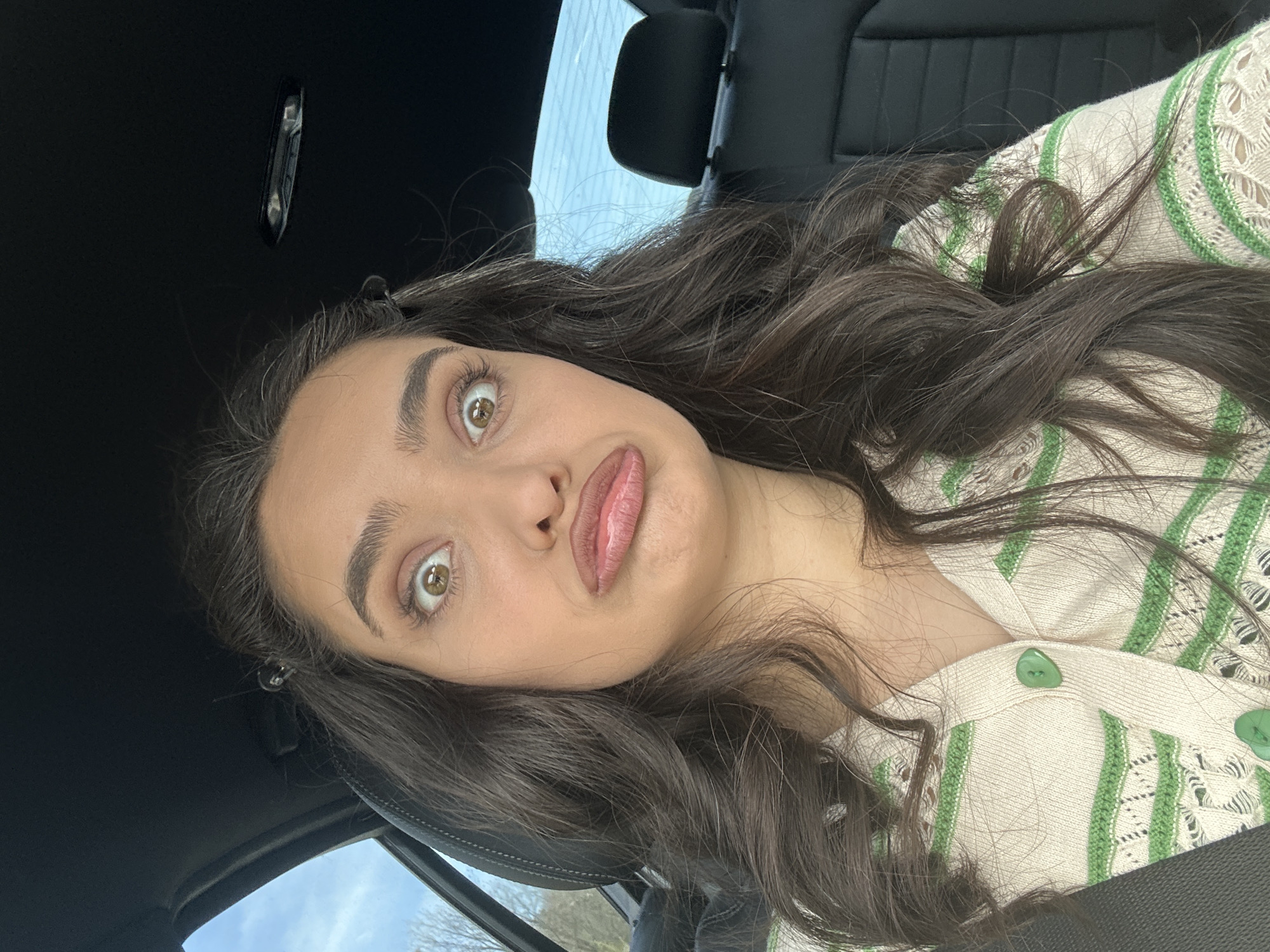 A young woman with long, wavy brown hair and light skin is in the driver's seat of a car. She is wearing a cream-colored sweater with green stripes and buttons. Her eyes are wide and her lips are puckered in a playful expression. The interior of the car is black. Through the car window, one can see a hint of sky and trees, suggesting an outdoor setting. It is impossible to discern the exact location in Zagorsko, Bulgaria, from this image.