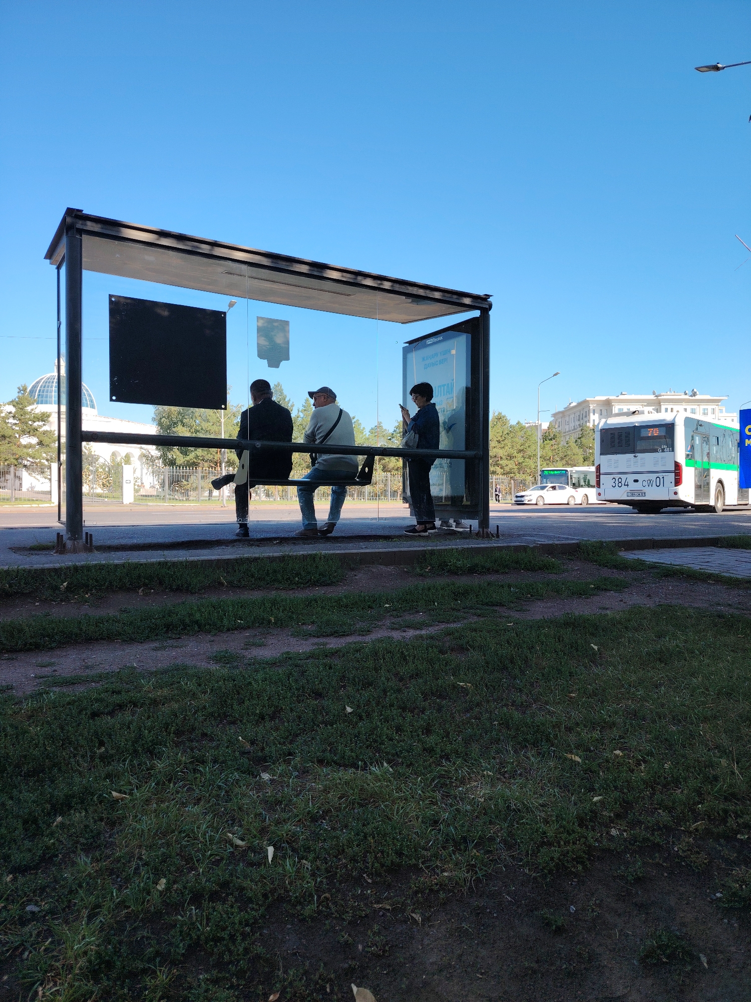 A bus stop in Astana, Kazakhstan, is shown with a clear blue sky. Three people are present at the bus stop. Two men are sitting on a bench inside the glass shelter, and one person, appearing to be a woman, is standing and looking at her phone. A white bus with green accents is visible in the background, along with other vehicles and buildings. The bus stop has a black bench and a glass enclosure with advertisements. The foreground is covered with grass.