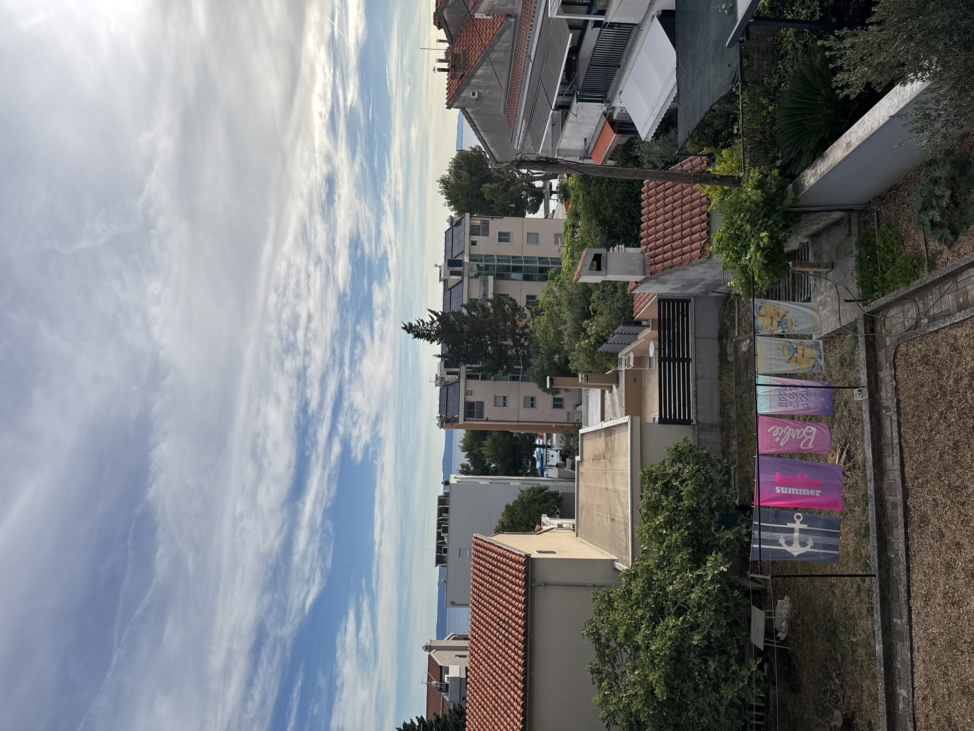 In Makarska, Croatia, a residential area is depicted with a view of the Adriatic Sea. Buildings with red-tiled roofs and apartment complexes are visible, some equipped with solar panels. The foreground features a clothesline with several colorful towels, including one with an anchor design, one that reads "hello summer," another with "Barbie," and two featuring characters from the animated movie "Despicable Me." Green trees and vegetation surround the buildings, and a portion of a garden is visible. The sky is partly cloudy with blue sky visible.