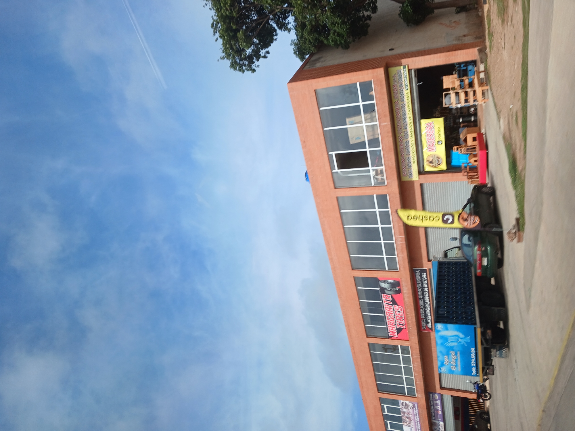 The image shows a commercial building in El Valle del Espíritu Santo, Venezuela, under a cloudy blue sky. The building is a single-story structure with an orange facade and large glass windows. Several businesses appear to be located here, indicated by signage. One prominent sign reads "MARGARITA TIRES". In front of the building, a blue truck with "El Angel" written on its side is parked. To the right of the truck, a green car is visible. Another sign, yellow with red lettering, displays "MADERA" and appears to be an advertisement for a carpentry business, with wooden furniture and structures displayed outside. A motorcycle is parked to the left of the truck. The ground in front of the building is paved, with some dirt and vegetation to the right. Trees are visible to the right of the building. A yellow flag with the word "cashea" is also present.