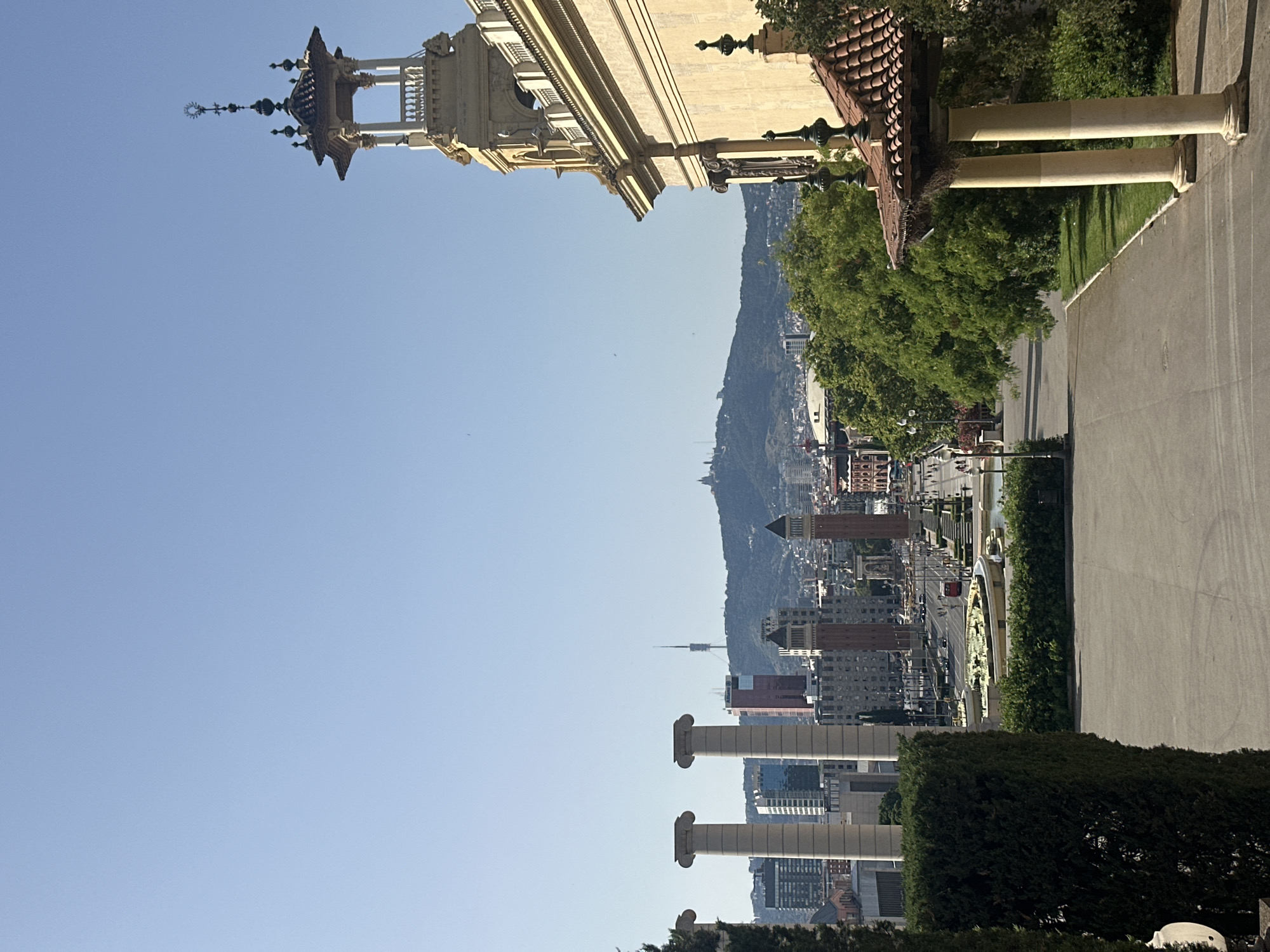 The image displays a scenic view of Barcelona, Spain, from a high vantage point. In the foreground, a paved pathway leads down towards a city vista. To the right, a light-yellow building with ornate architectural details, including columns and a tiled roof, dominates the scene. Adjacent to this building, a verdant lawn and lush green trees create a natural buffer.
Further in the midground, a row of four large, cream-colored columns stands, leading the eye towards the cityscape. Beyond these columns, the urban landscape of Barcelona unfolds. Several tall buildings, including modern skyscrapers and older, red-brick structures, are visible. Two prominent cylindrical towers with red brick bases and darker tops stand out in the center of the frame, framing a fountain with surrounding greenery.
In the background, a hazy mountain range stretches across the horizon. A tall, slender telecommunications tower is perched atop one of the hills, and another distant structure, possibly a church or monument, is visible on a higher peak. The sky is a clear, pale blue, indicating a bright, sunny day. The overall impression is one of a grand, urban panorama viewed from a privileged position within the city.