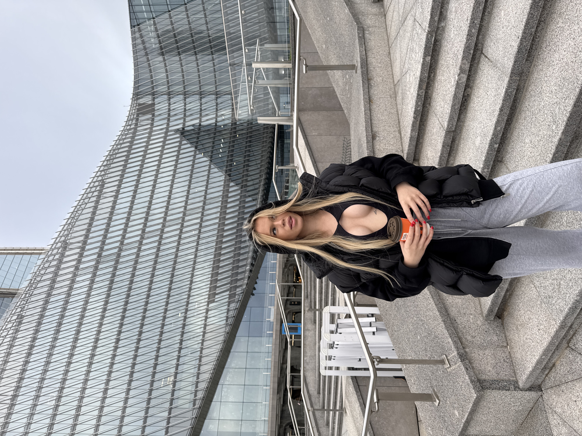 The image shows a young woman with blonde hair sitting on a set of granite stairs. She is wearing a black puffer jacket, a black sports bra, and grey sweatpants. She is holding an orange coffee cup in her right hand. Behind her, a modern glass-fronted building with a curved facade rises into a grey, overcast sky. The architecture suggests an urban environment. The woman's gaze is directed towards the camera, and her expression is neutral. The lighting and the overcast sky suggest it is daytime, likely with cool weather, given her clothing. The context provided places the scene in Saint Petersburg, Russia, and the modern architecture of the building aligns with contemporary urban development. There is no visible text in the image.