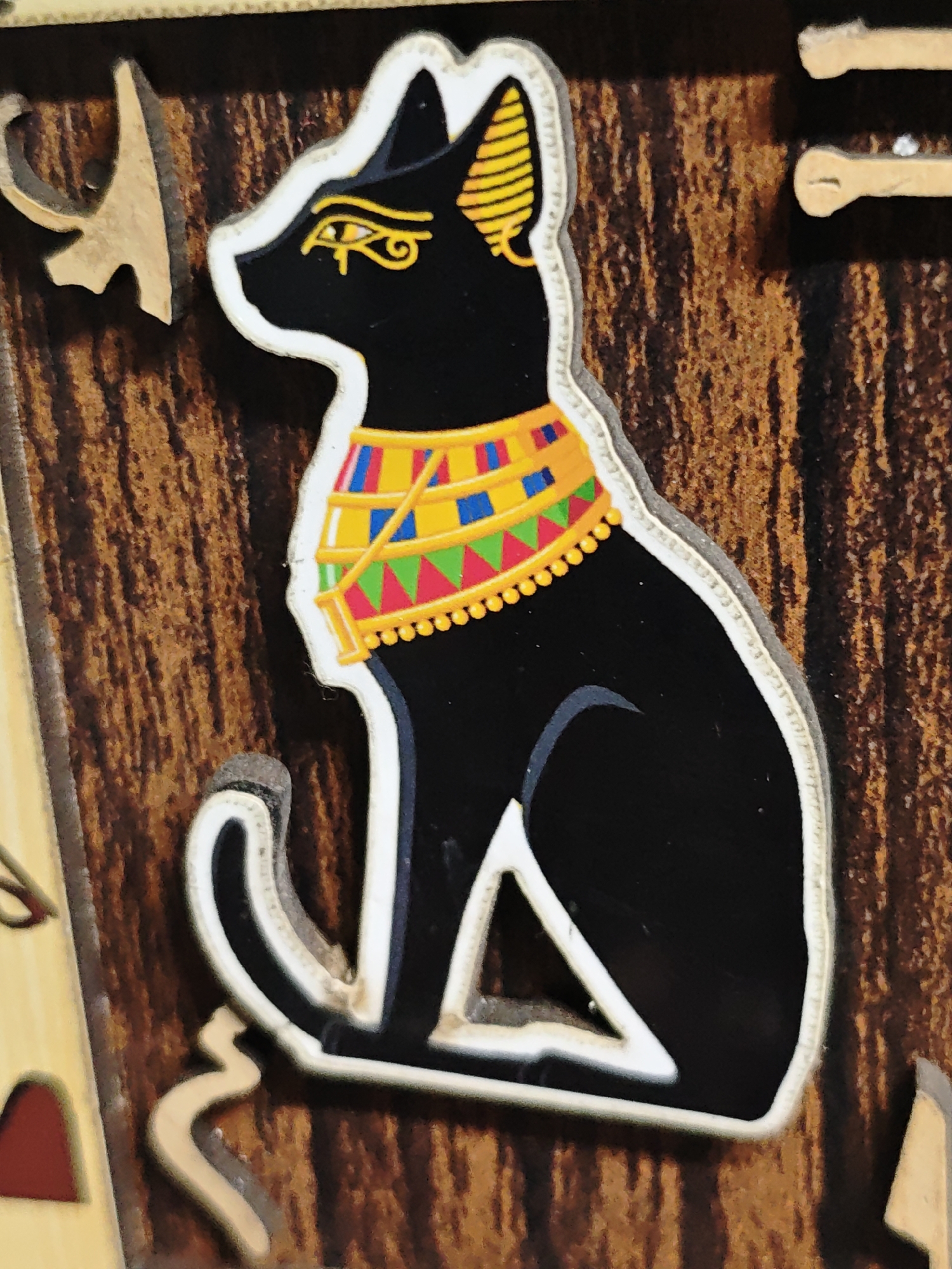A close-up photograph depicts a stylized, Egyptian-themed black cat figure attached to a wooden surface. The cat is outlined in white, and decorated with a colorful collar of yellow, blue, green, and red triangles and beads. It has an Egyptian eye symbol on its face and yellow striped markings on its ears. The wooden surface has a textured, brown and tan pattern. The image captures part of a larger object. No specific location in Боровково, Russia, can be determined from the image.