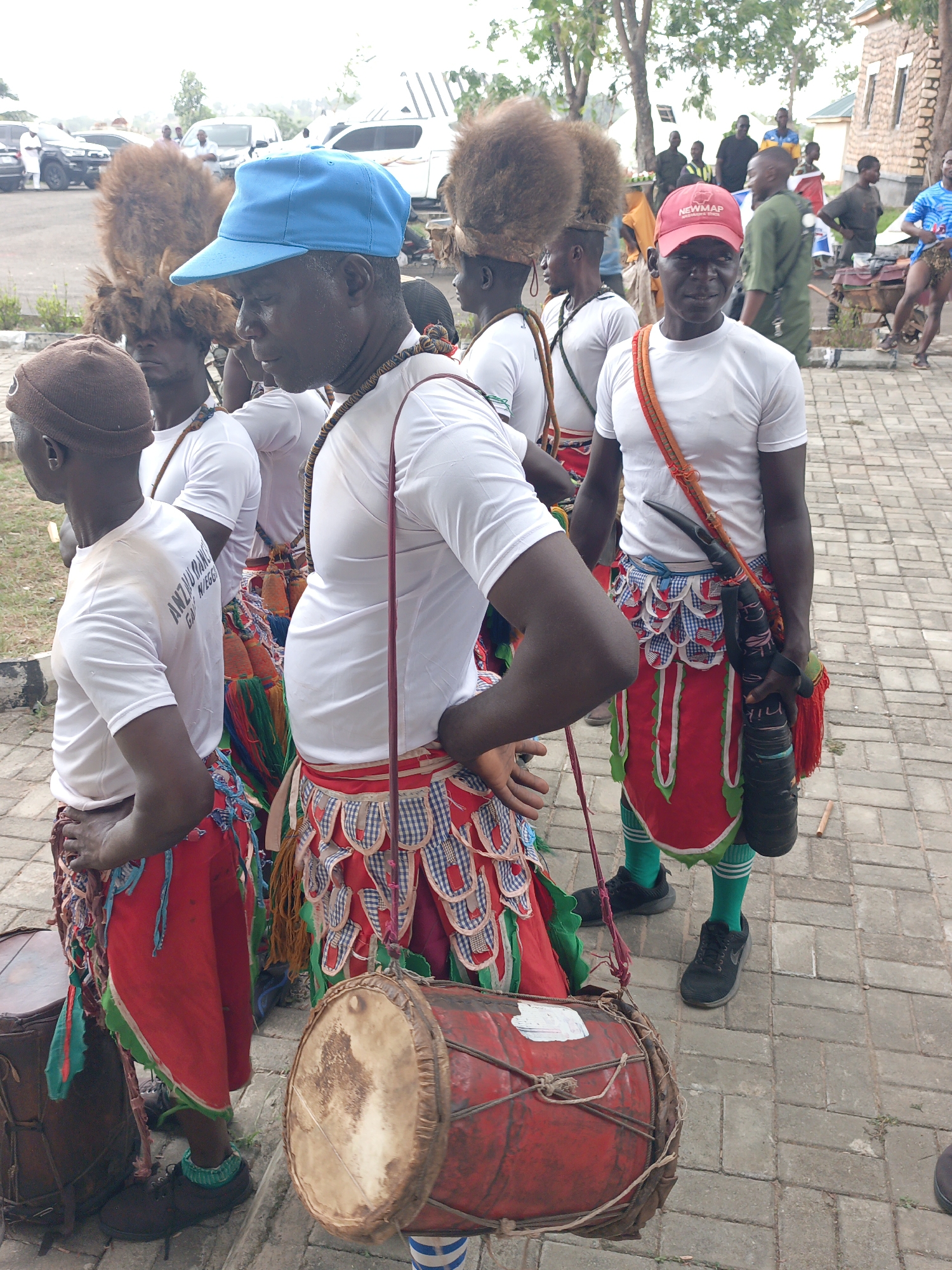 In Akwanga, Nigeria, a group of men are dressed in traditional attire, including white shirts, colorful skirts with fringe and patterns, and headdresses with large, furry extensions. Some are wearing caps: one blue and one red with white lettering. A man in the foreground carries a drum. Another man has a long, cylindrical object with a red tassel strapped across his body. Other people are visible in the background along with cars and buildings. The group is standing on a paved area.