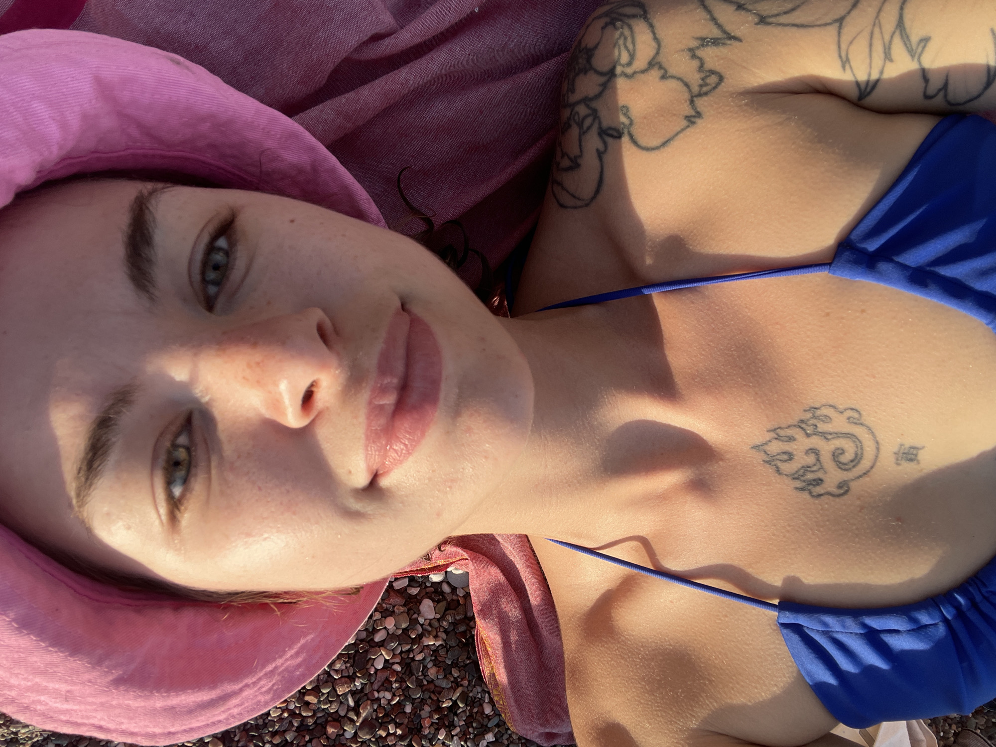 A woman lies on a beach in Rafailovići, Montenegro. She wears a pink bucket hat and a blue bikini top. Her face, with freckles, is visible, and she is smiling slightly. Tattoos are visible on her chest and upper arm. The beach is pebbled, and a pink scarf or blanket is also present. The lighting suggests it is daytime, possibly late afternoon due to the angle of the sun.