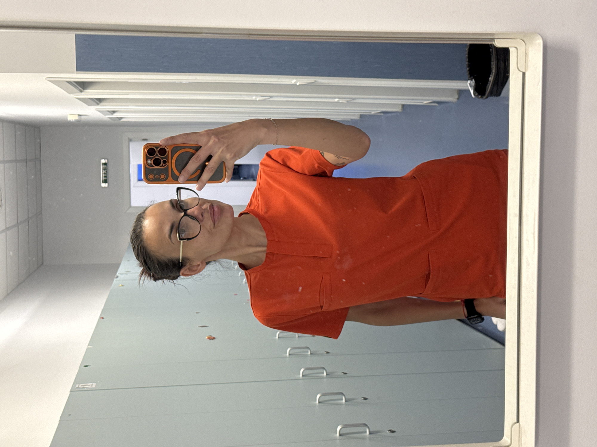 A woman is taking a mirror selfie in what appears to be a locker room. She is wearing an orange scrub top, black-rimmed glasses, and a wrist watch. She is holding an orange-cased cell phone. The background includes a row of light gray lockers, a hallway with a blue accent stripe, and a shoe sitting above. There are no identifying markers to confirm the location in Pleven, Bulgaria.