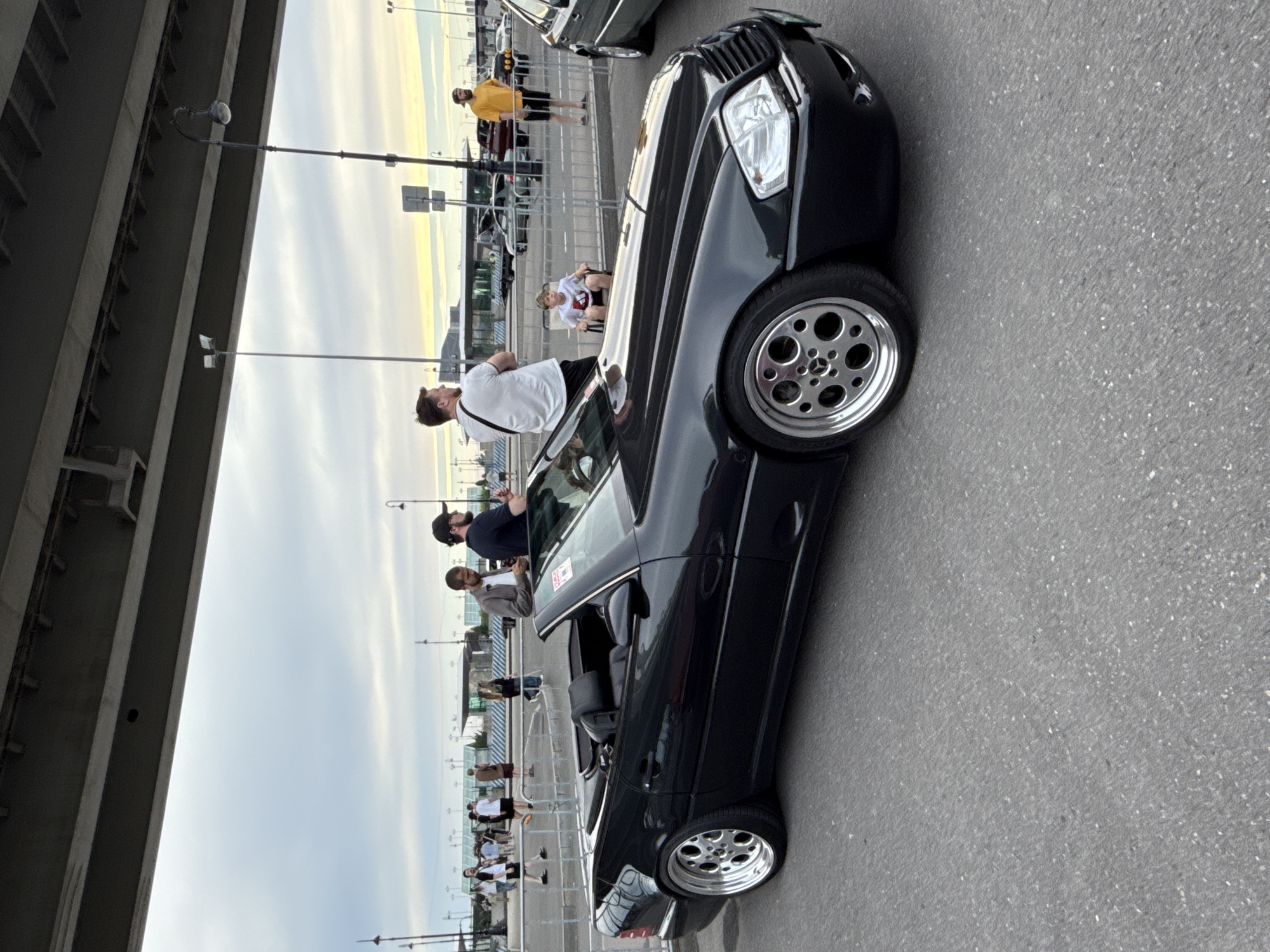 In Saint Petersburg, Russia, a black Mercedes-Benz SL convertible is parked on asphalt under a large overpass. Several people are gathered around the car and in the background. A young boy sits in a chair near the car. The sky is overcast with hints of yellow and orange near the horizon. The scene appears to be an outdoor event or gathering.
