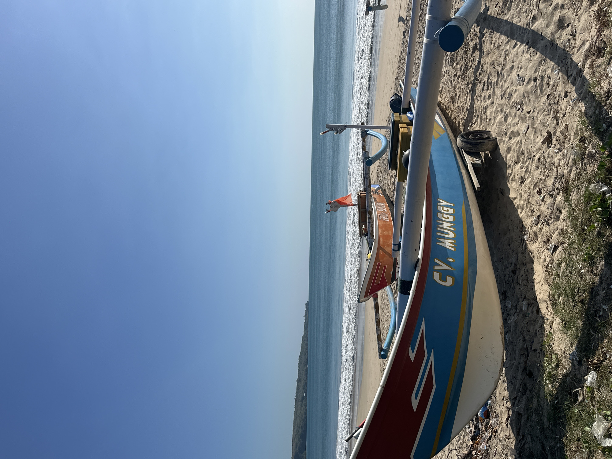 This is a ground-level, full-frame shot of a beach in Kuta, Indonesia, on a clear, sunny day. The foreground is dominated by the side of a blue and white fishing boat, with red and yellow trim. The boat has "CV. MUNGGY" written in white lettering on its side. It is resting on sandy ground, with some patches of green vegetation and scattered debris. A wheel is visible under the boat, suggesting it is on a trailer. Behind this boat, partially visible, is another fishing boat that is orange and white. The boats are positioned on a sandy beach. The middle ground features the ocean, with gentle waves rolling onto the shore. The water is a serene blue, transitioning to a lighter shade near the shore. A distant headland, covered in lush green vegetation, forms the horizon line in the background, meeting a clear, bright blue sky. The overall scene conveys a sense of tranquility and coastal life.