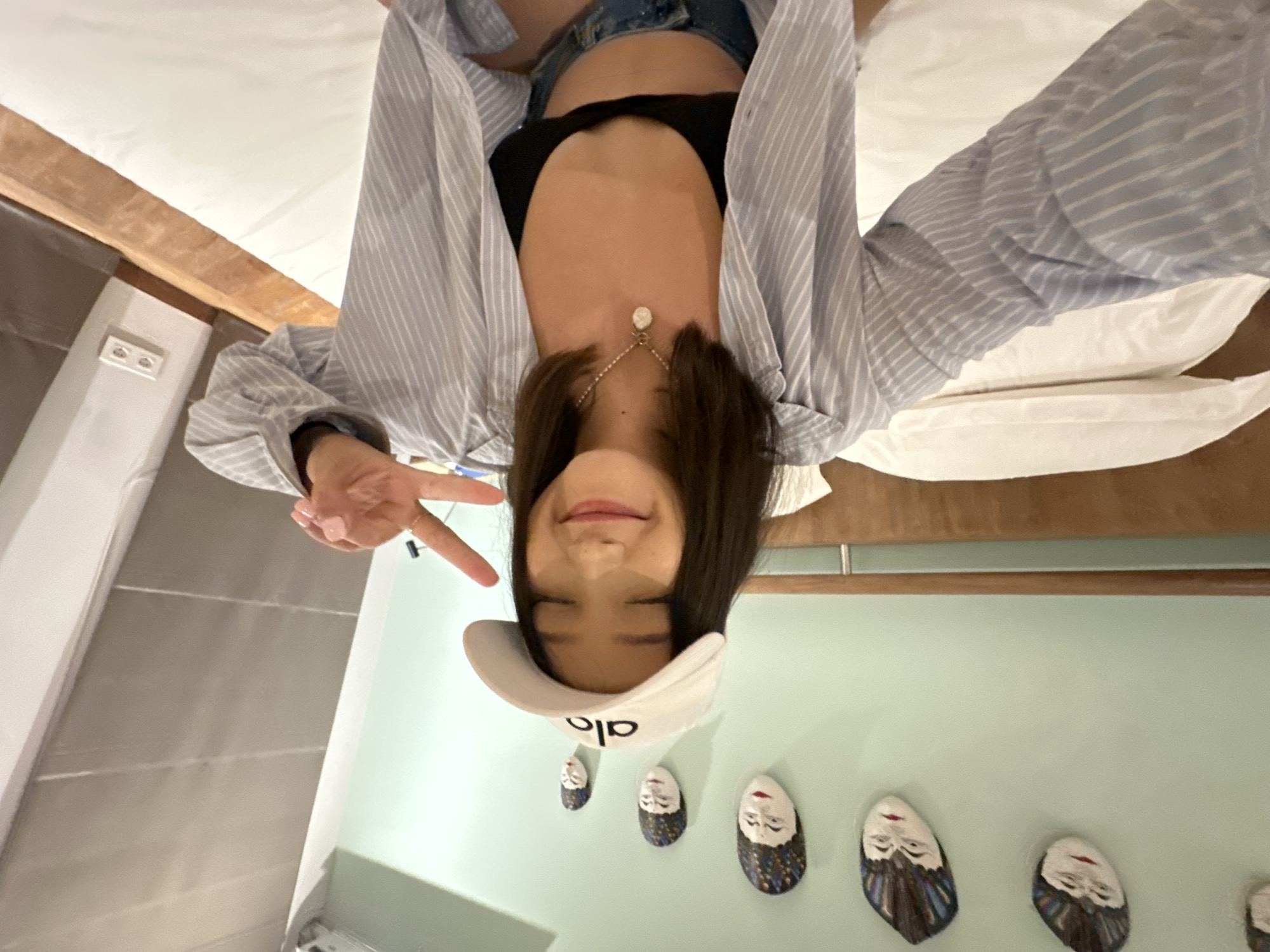 A young woman with dark hair, wearing a white baseball cap, a light blue and white striped unbuttoned shirt over a black bralette, and denim shorts, poses for a selfie on a bed in a room. She has her eyes closed and is smiling, holding up two fingers in a peace sign. A necklace with a light-colored pendant is visible. The room has a light green wall with several decorative masks hanging on it, and a wooden headboard. The bed is made with white linens. The scene appears to be in a bedroom in North Kuta, Indonesia, as indicated by the decor and the overall ambiance.