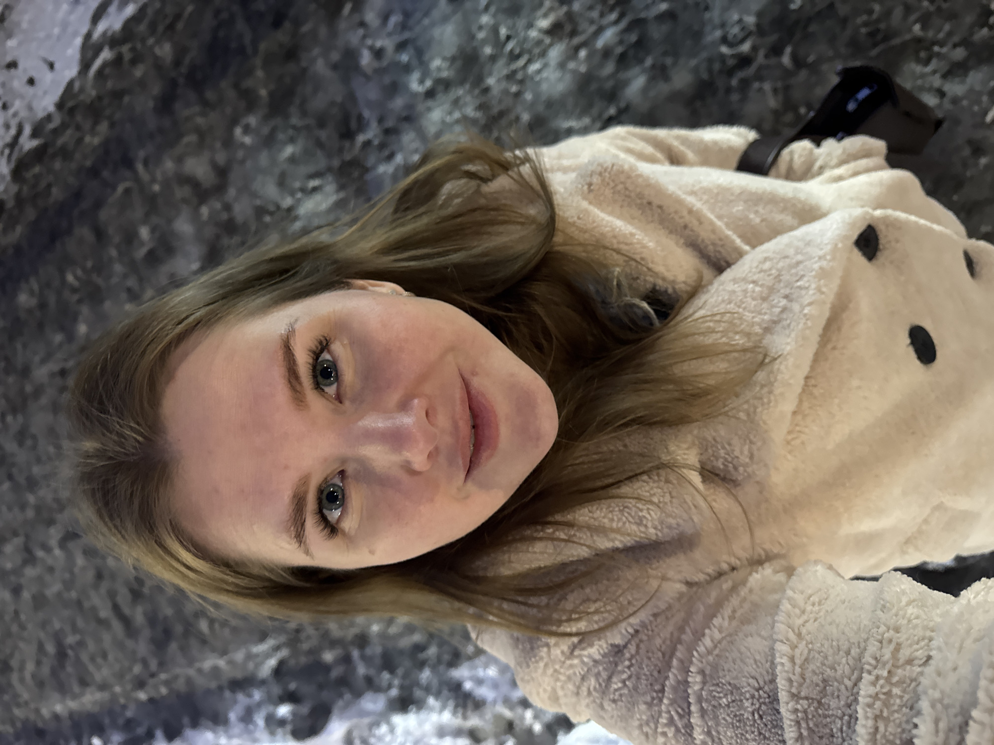 A young adult woman is featured in a close-up, upward-angled selfie, set against a dark, textured background. She has light skin, blue-grey eyes, and long, light brown hair that frames her face. A faint, pleasant smile plays on her lips, and her gaze is directed towards the camera. She is wearing a thick, warm, cream or beige-colored fuzzy/fleece top, possibly a hoodie, adorned with dark, circular patterns, resembling spots or polka dots, visible on her right side. A black strap or part of a bag is visible behind her right shoulder. The background appears to be a rugged, dark surface, possibly resembling rock or ice, suggesting an environment like a cave or a cold, natural or artificial installation. Given the location context of Perm, Russia, this setting could be an ice cave or a similar cold-weather attraction. The warm clothing further reinforces the inference of a chilly environment. No text is visible in the image.