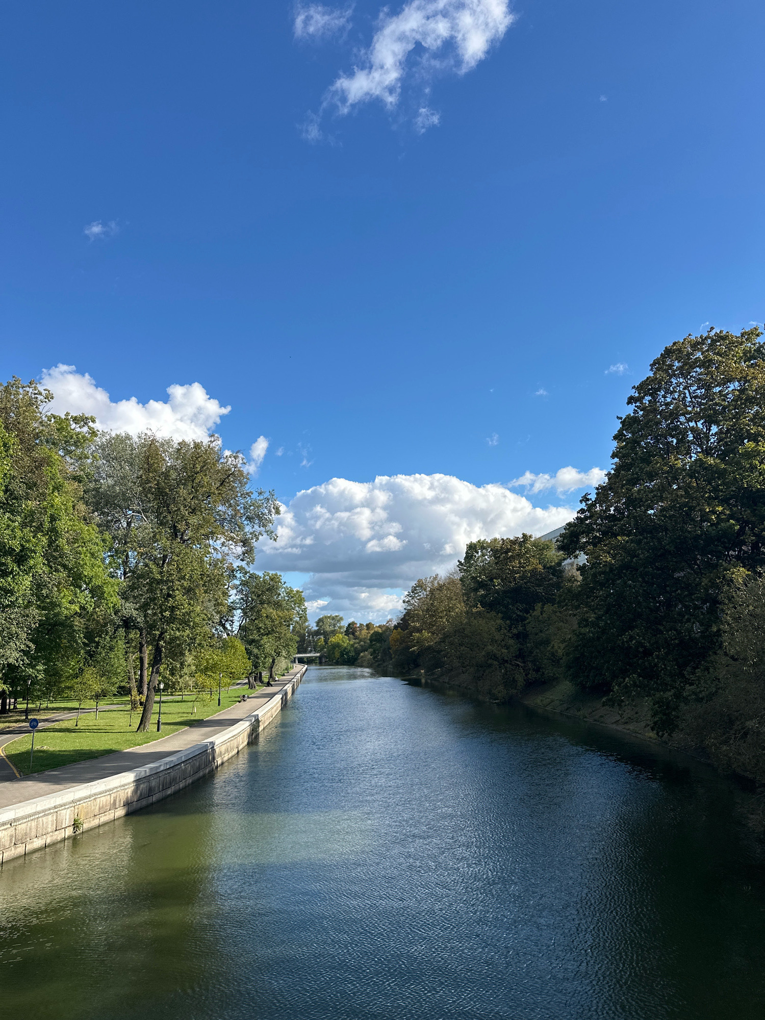 The image depicts a wide canal in the city of Minsk, Belarus, likely within the Svislach River embankment area, given the characteristic architecture and greenery. The canal is flanked by well-maintained concrete embankments and lush green parkland on the left, with mature trees lining the pathway. On the right bank, a dense cluster of trees forms a natural barrier. The water in the canal reflects the bright blue sky, which is dotted with scattered white clouds. A paved walkway runs along the left bank, and in the distance, a bridge is visible, suggesting the canal continues further into the city. The scene is tranquil, with no visible people or activity, indicating a peaceful urban environment. The overall impression is one of a scenic and well-preserved public space in Minsk.